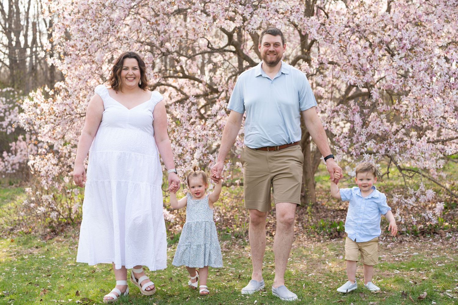 A family wearing coordinated white and blue outfits poses together under blooming cherry blossom trees in spring.