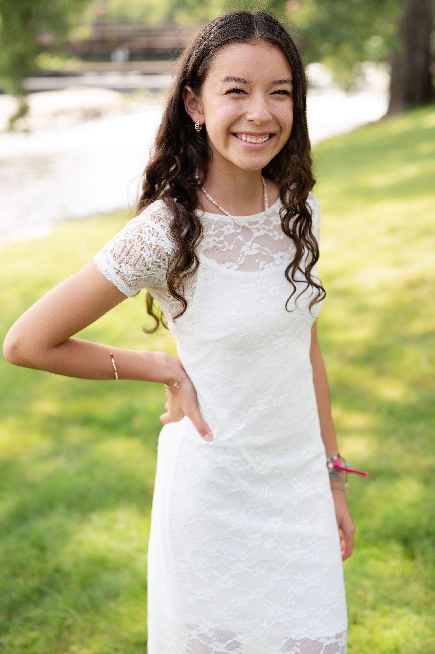 A person in a white lace dress poses playfully outdoors on a sunny day with a grassy background.