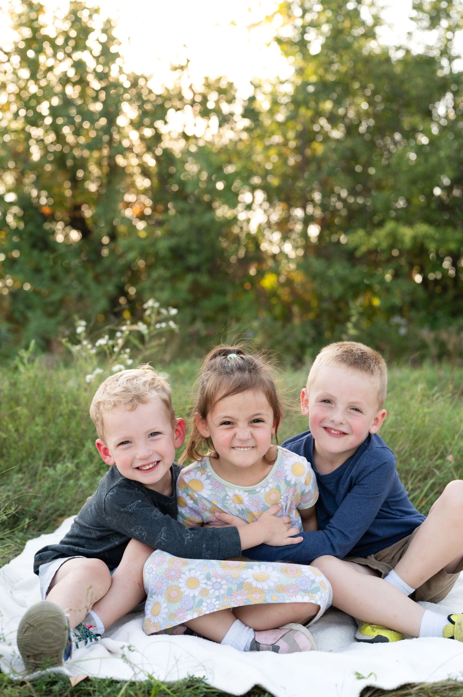 Three young children lying on a white blanket outdoors smiling and laughing together in a grassy setting at sunset.