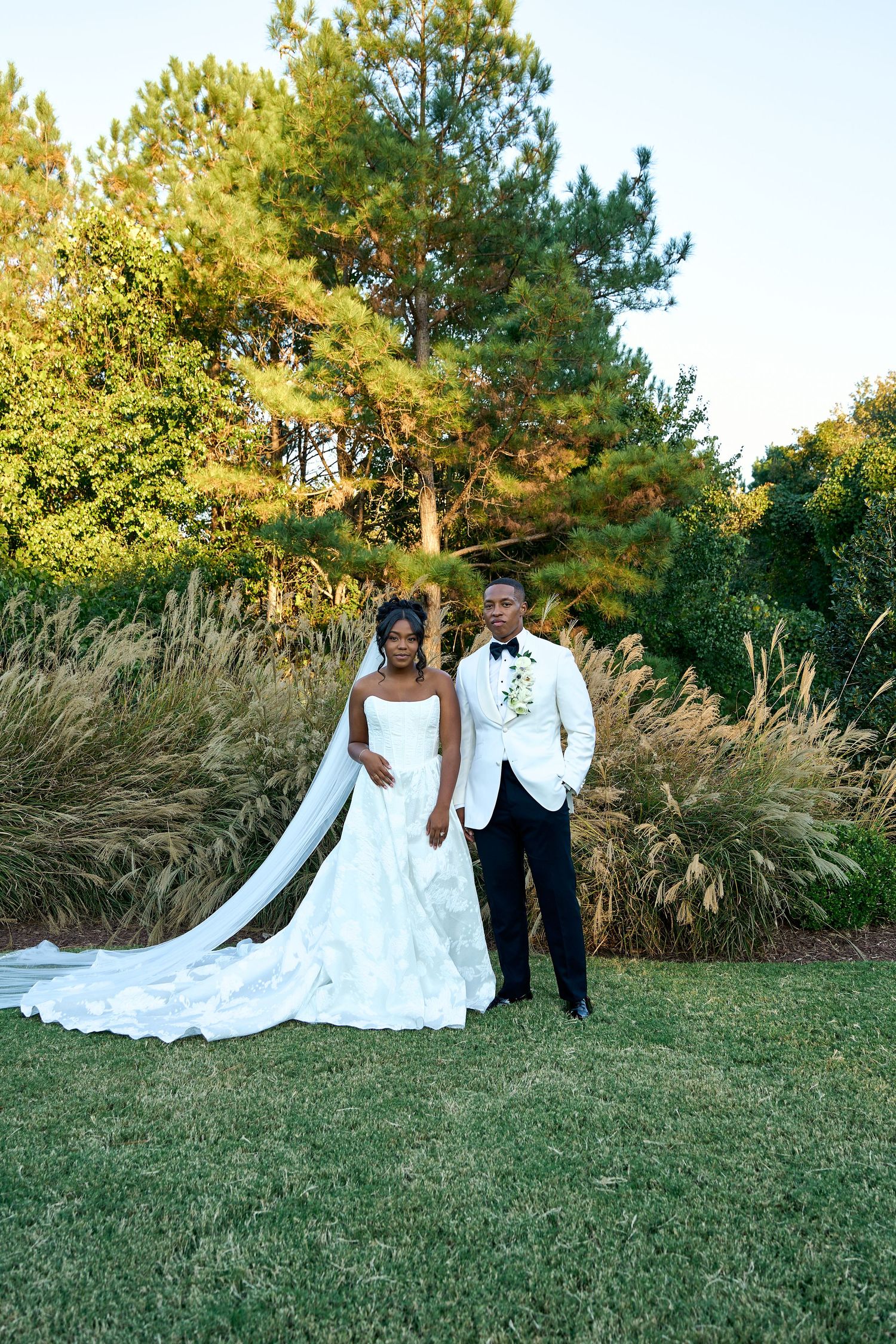 Bride in white gown and groom in white tuxedo jacket pose together on green lawn with tall trees backdrop.