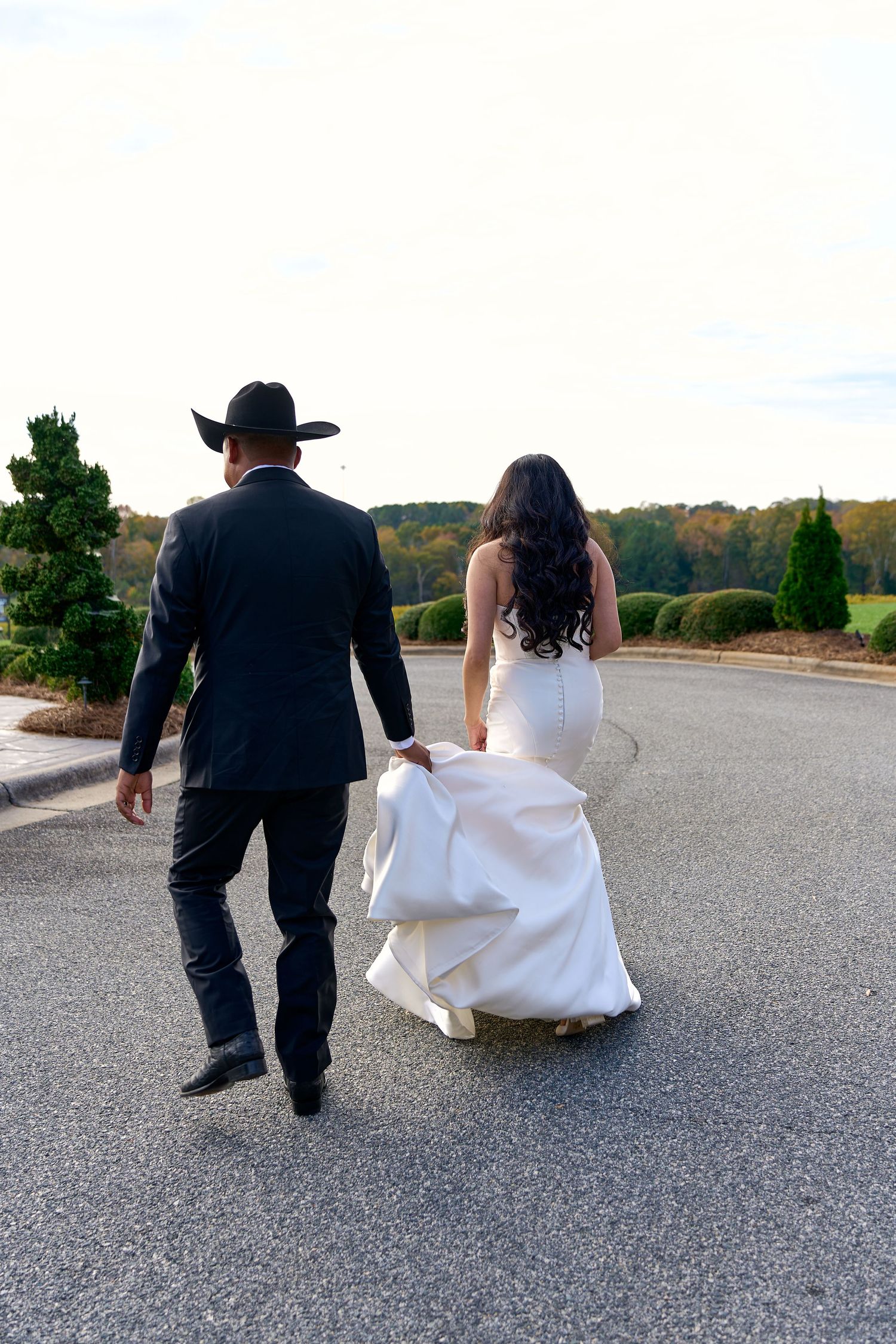 A bride in a white gown and groom in a black suit and cowboy hat walk together on a paved path outdoors.
