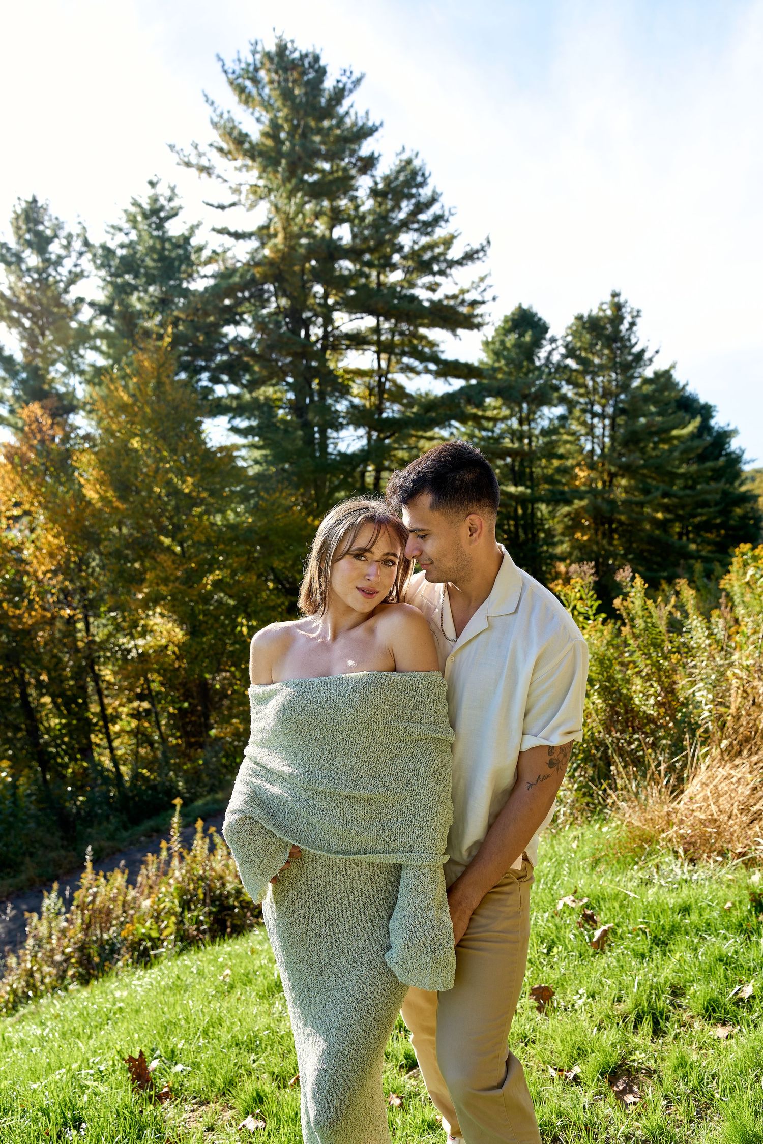 A couple embraces outdoors in autumn, surrounded by tall evergreen trees and golden foliage on a sunny day.