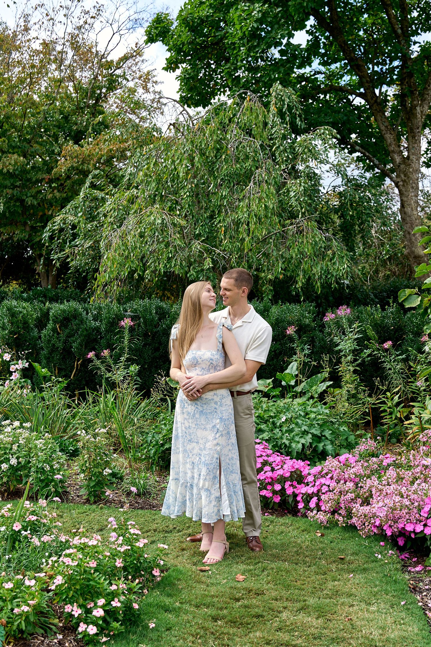 A couple embraces in a lush garden surrounded by pink flowers and green trees on a bright day.