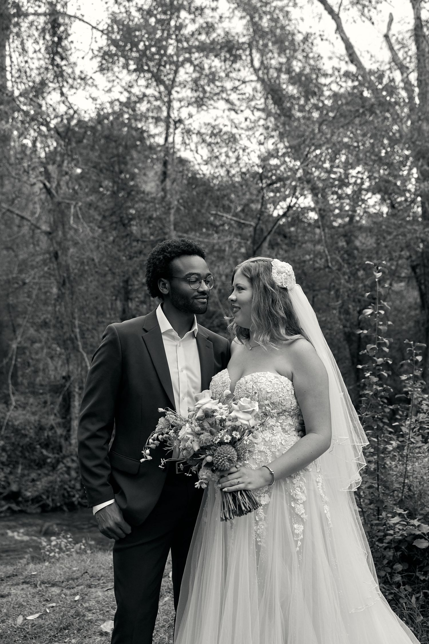 Black and white photo of an interracial couple on their wedding day, bride in lace gown holding bouquet, groom in suit, outdoors.