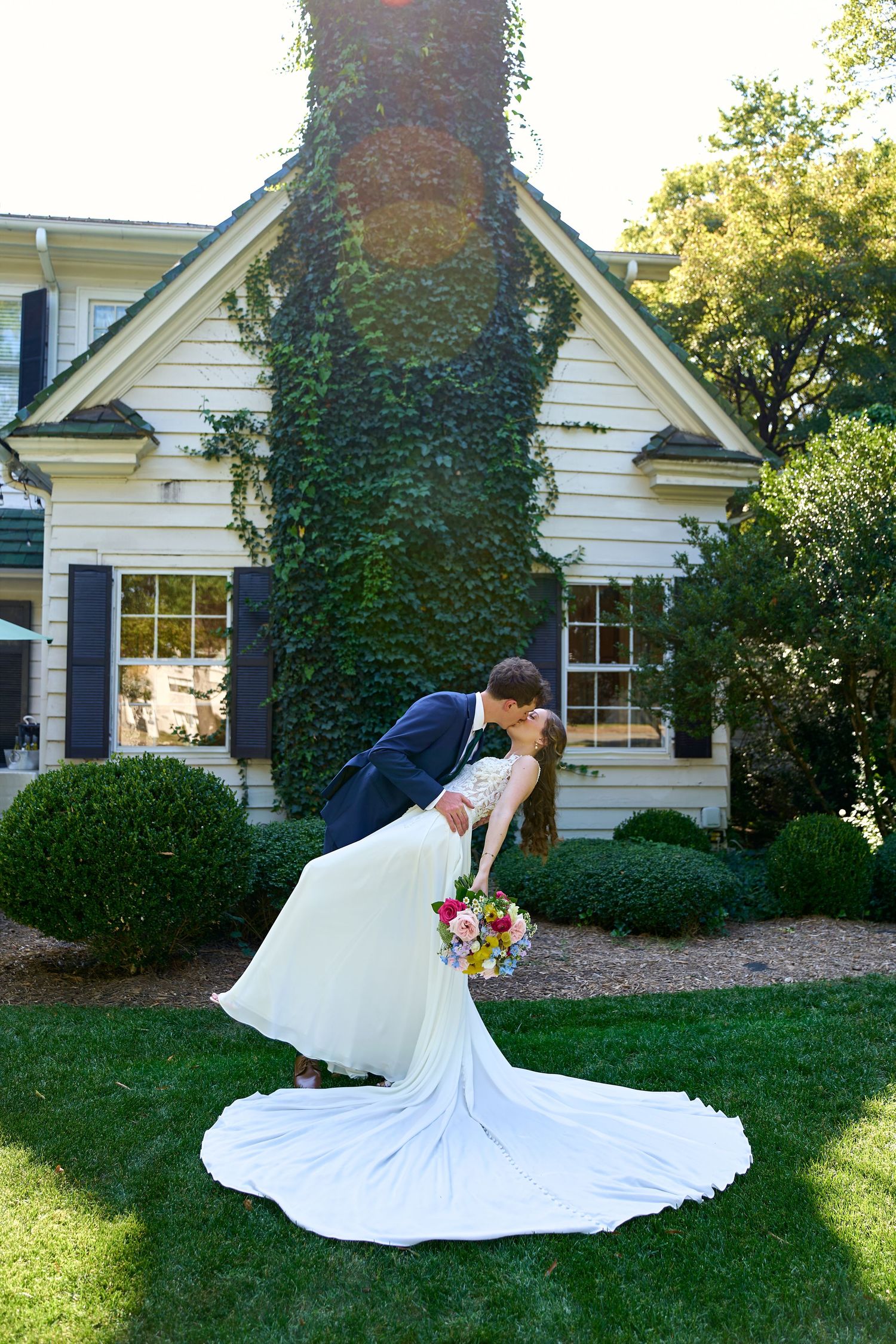 A groom dips his bride for a romantic kiss in front of an ivy-covered white house on their wedding day.