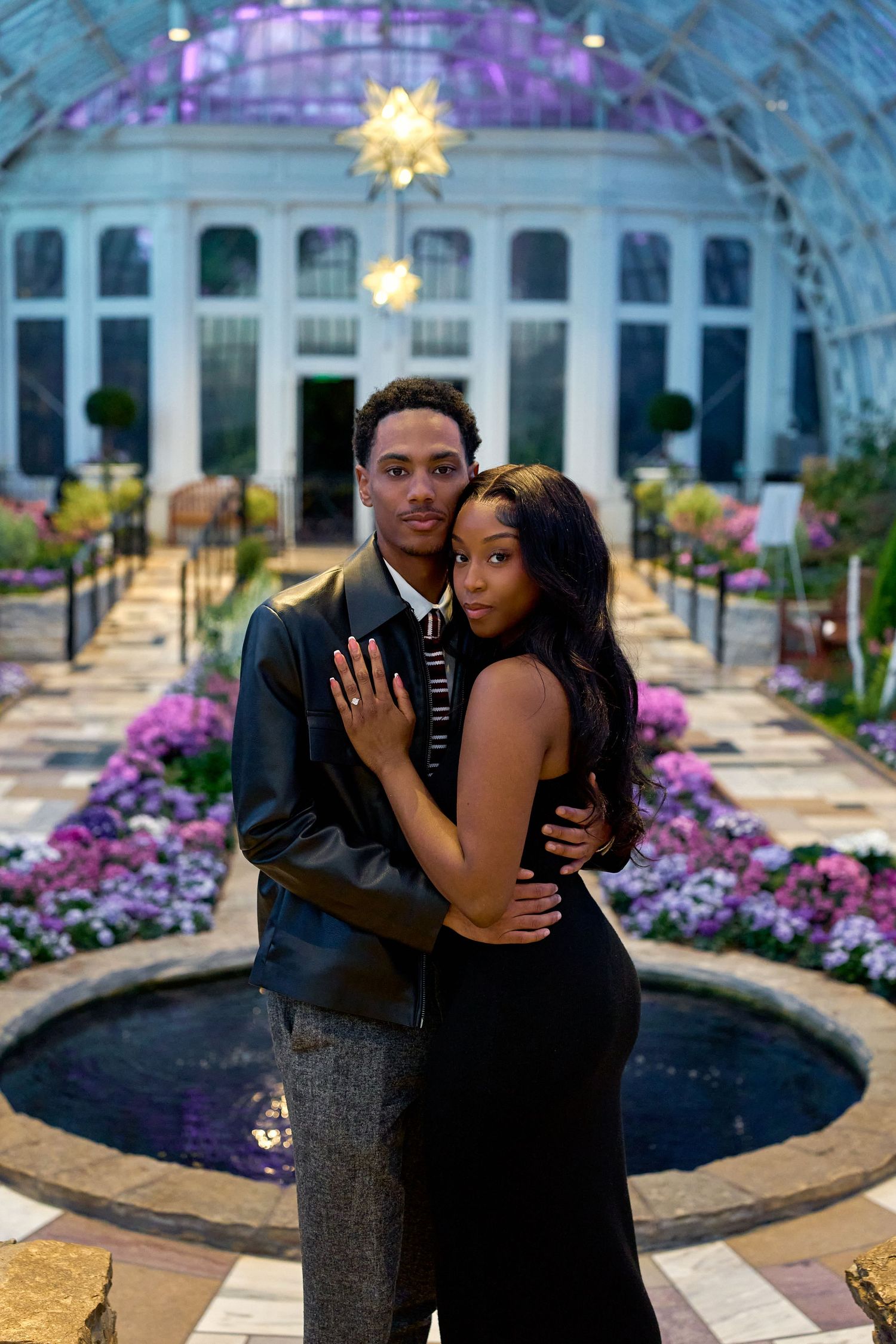 Elegant couple posing in a stunning greenhouse conservatory with purple flowers and a fountain backdrop.