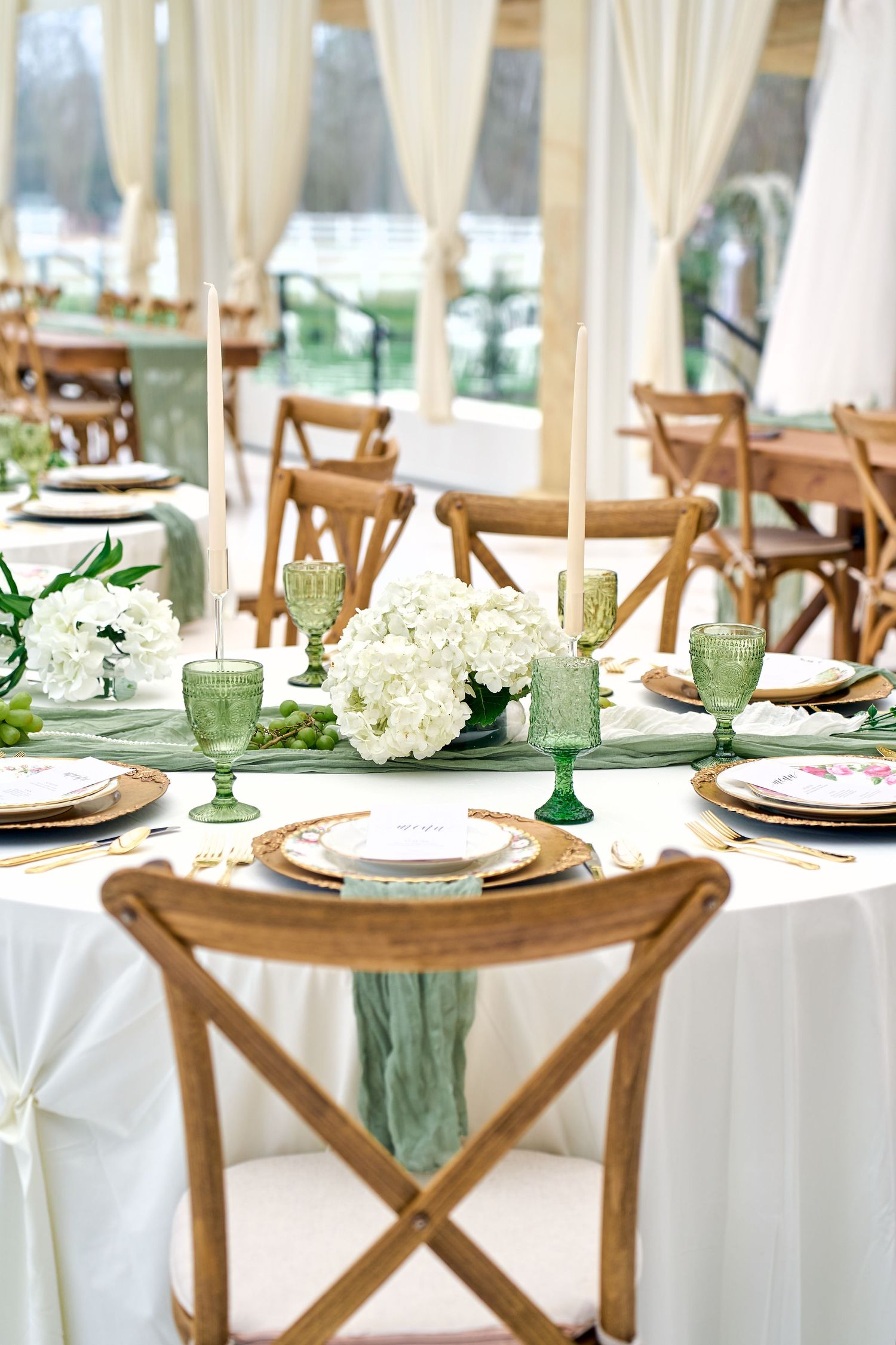 Elegant wedding table setting with white hydrangeas, green glassware, and rustic wooden cross-back chairs.