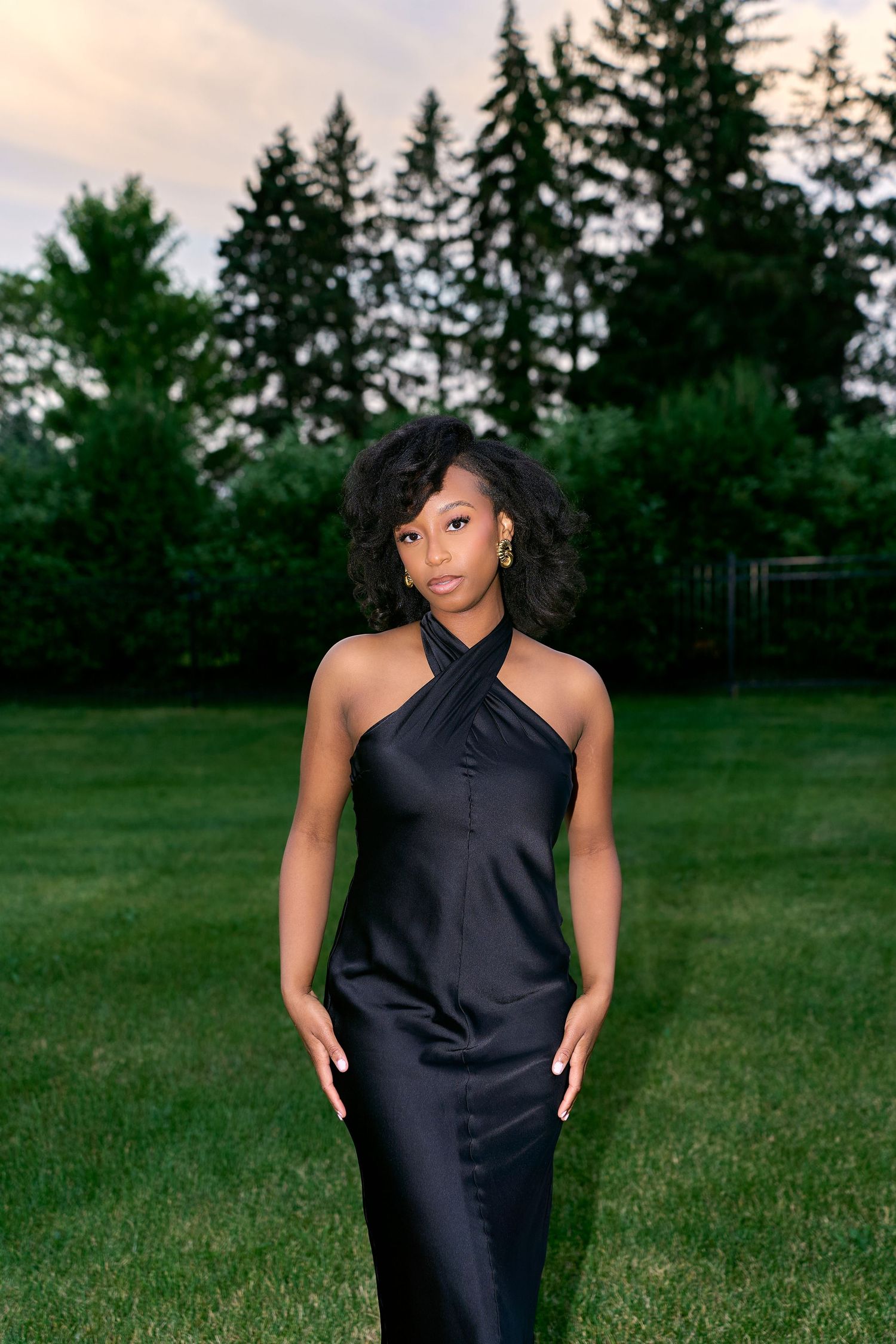 A person poses in a sleek black sleeveless outfit against a green grassy field with evergreen trees in the background.