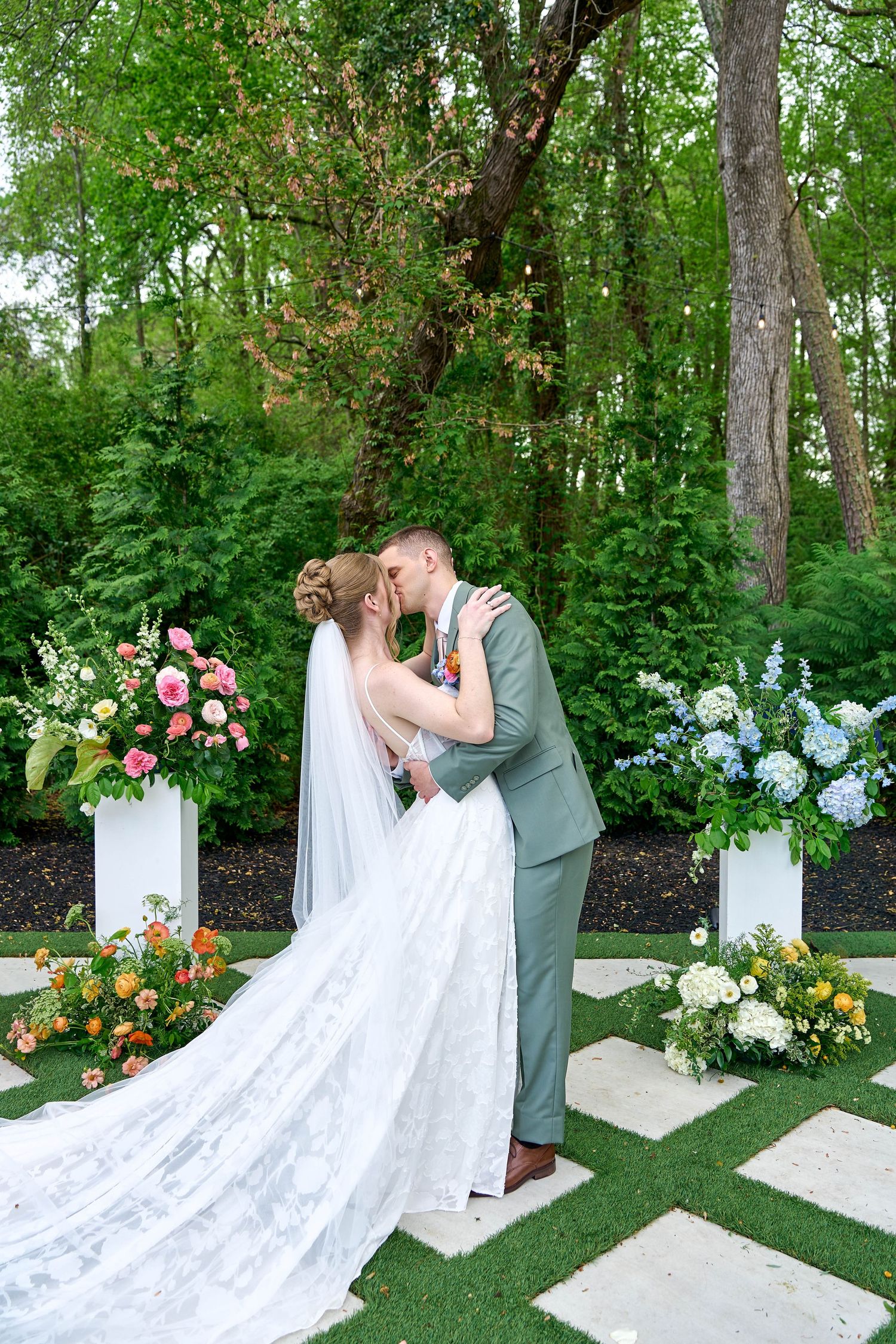 Bride and groom share first kiss at outdoor garden wedding ceremony surrounded by colorful floral arrangements.