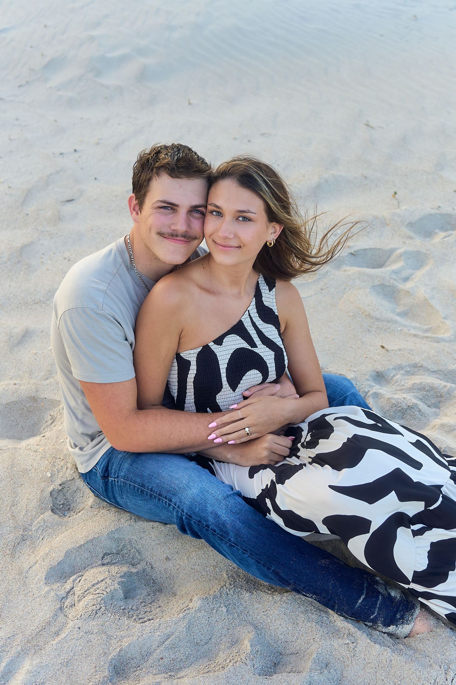 A couple relaxes together on sandy beach, with her wearing a black and white patterned dress.