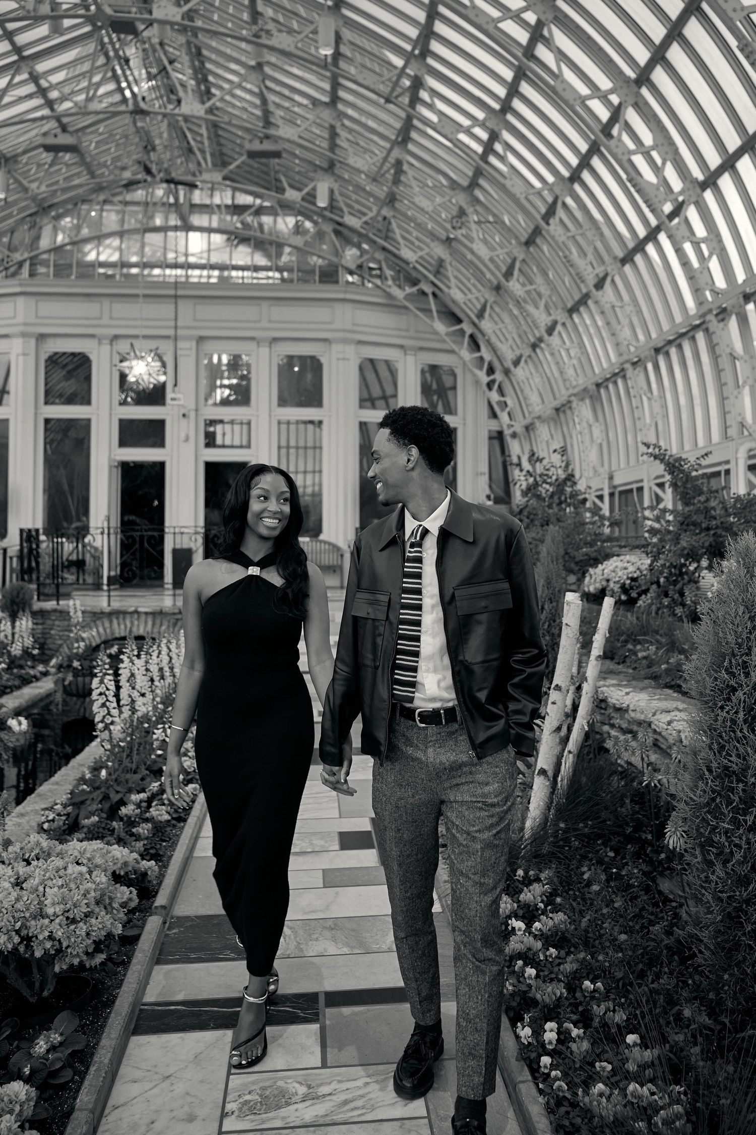 A couple walks hand-in-hand through a stunning glass greenhouse conservatory surrounded by lush flowers.