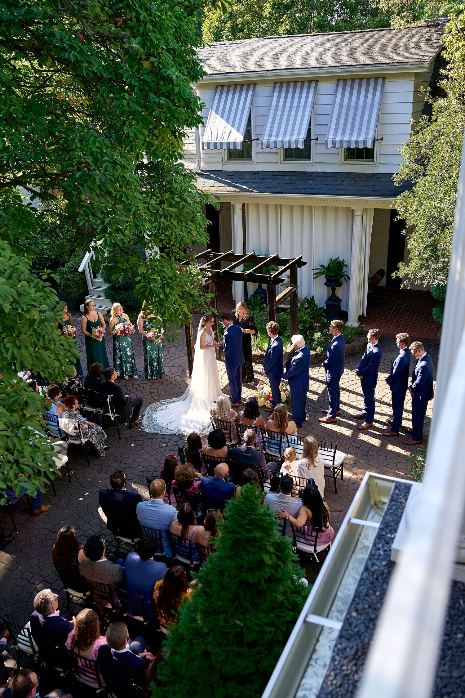 Aerial view of an elegant outdoor wedding ceremony in a garden courtyard with bridesmaids, groomsmen, and seated guests.