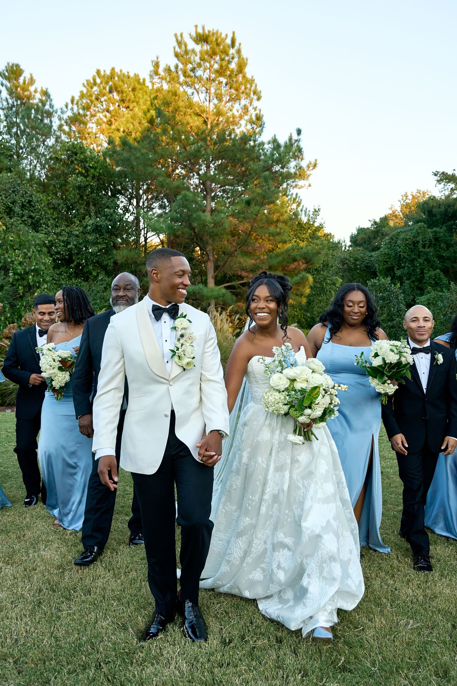 A joyful bride and groom walk hand-in-hand outdoors, surrounded by their wedding party in blue gowns and black tuxedos.