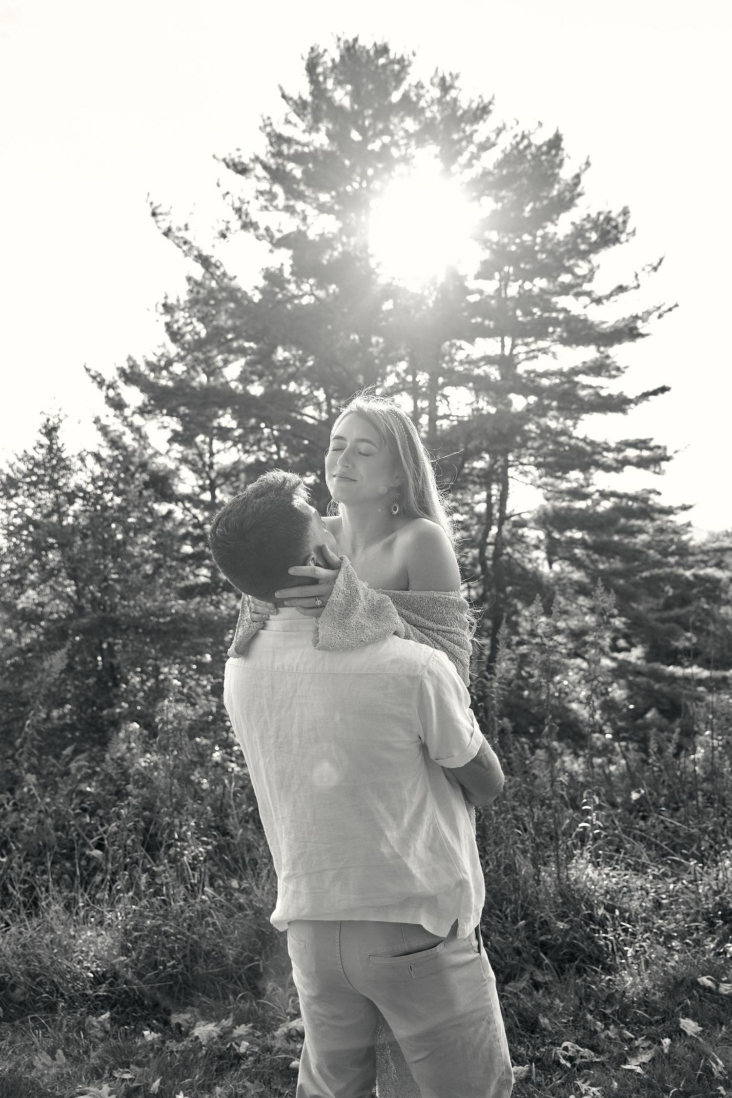 Black and white photo of a man lifting a woman in a sunlit forest, sharing a romantic moment among tall pine trees.