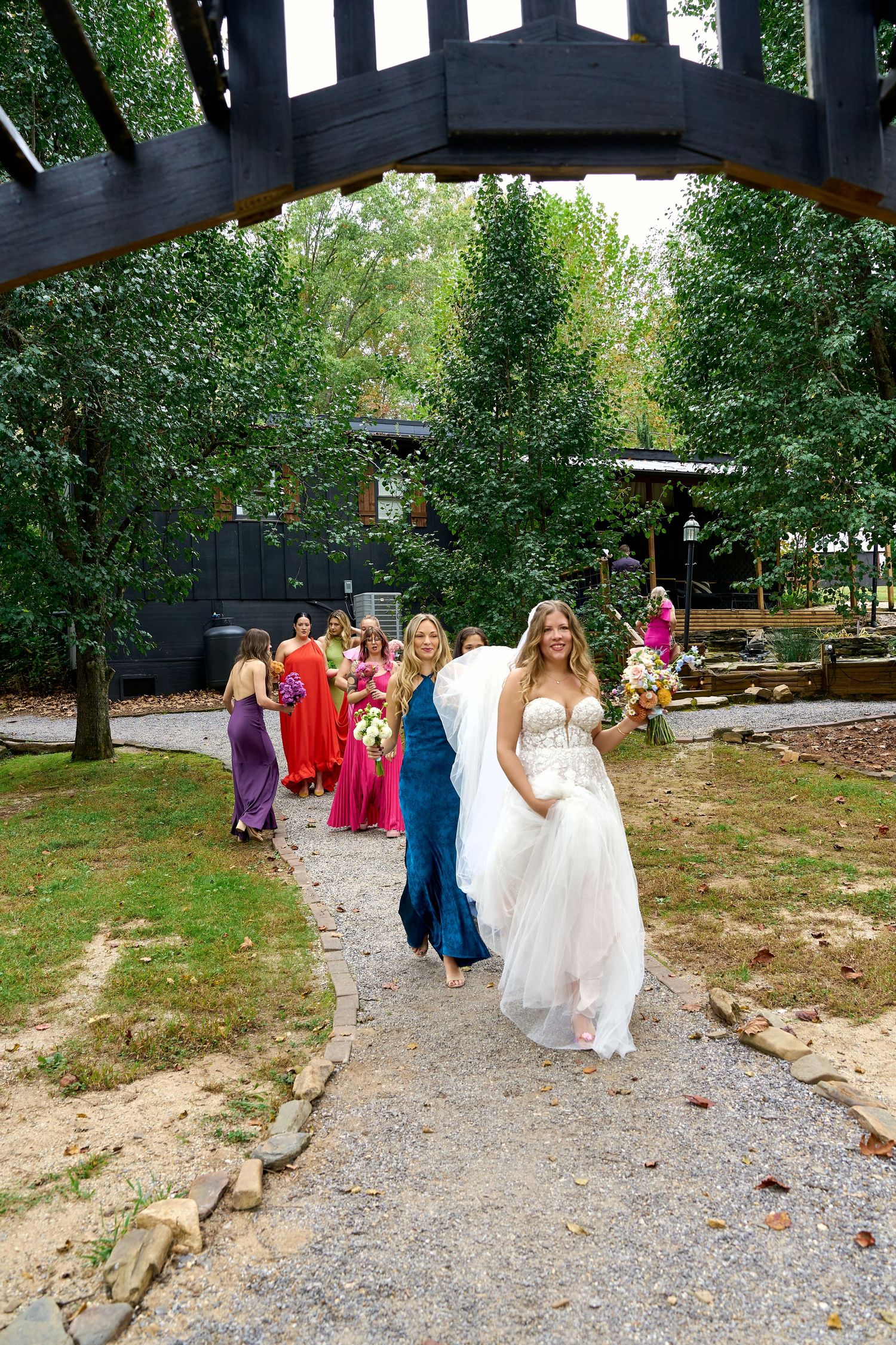 Bride in white gown leads colorful bridesmaids down garden path at outdoor wedding venue surrounded by trees.