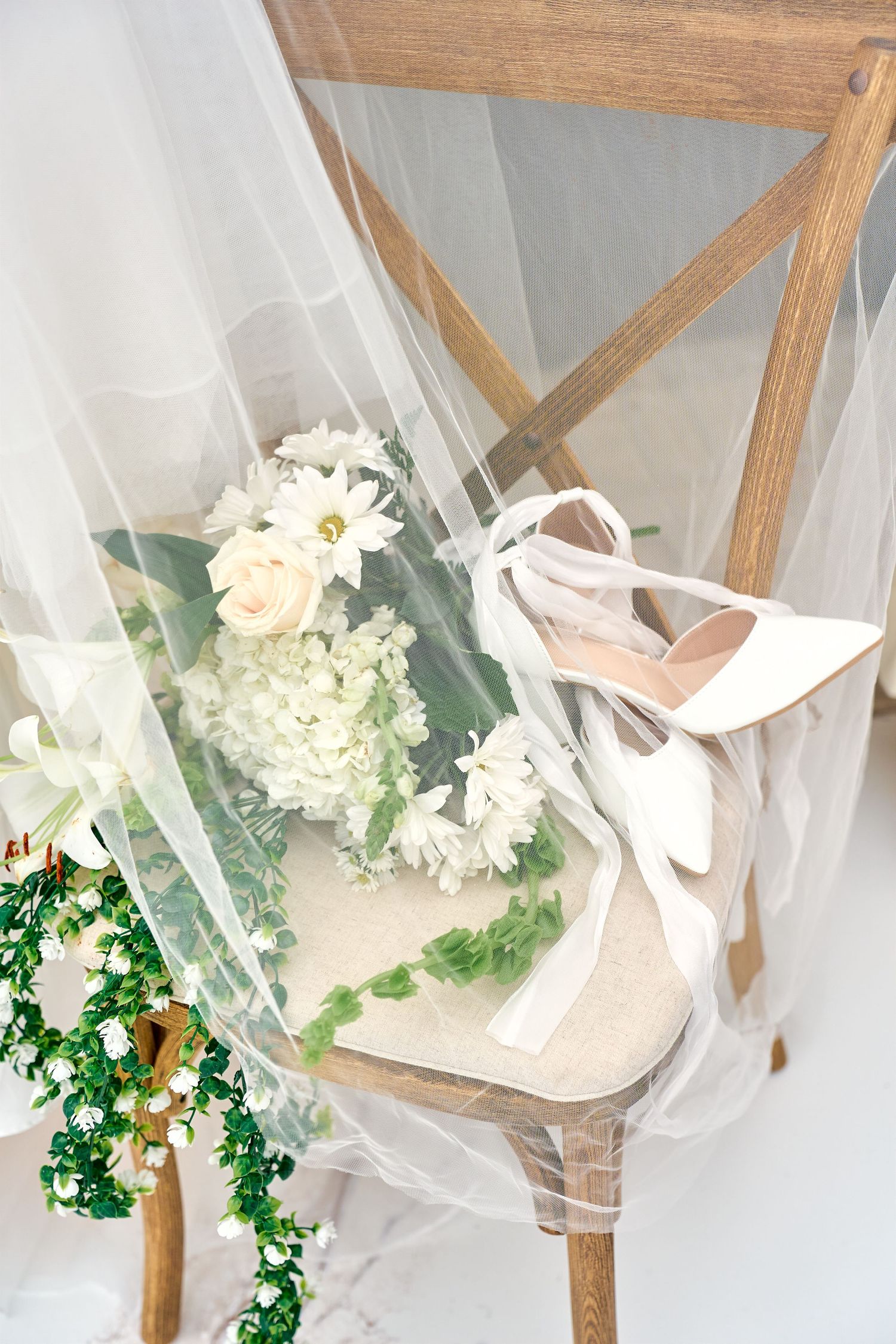 White bridal high heels and a delicate bouquet with roses and hydrangeas rest on a wooden chair draped with tulle fabric.