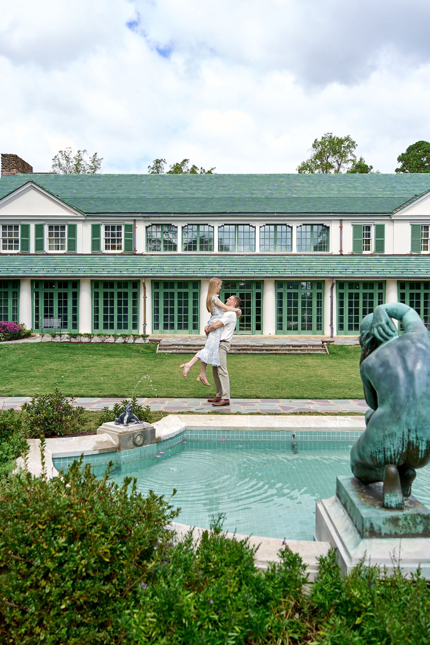 Couple embracing by a pool in front of an elegant green-roofed estate with manicured gardens and a bronze lion statue.