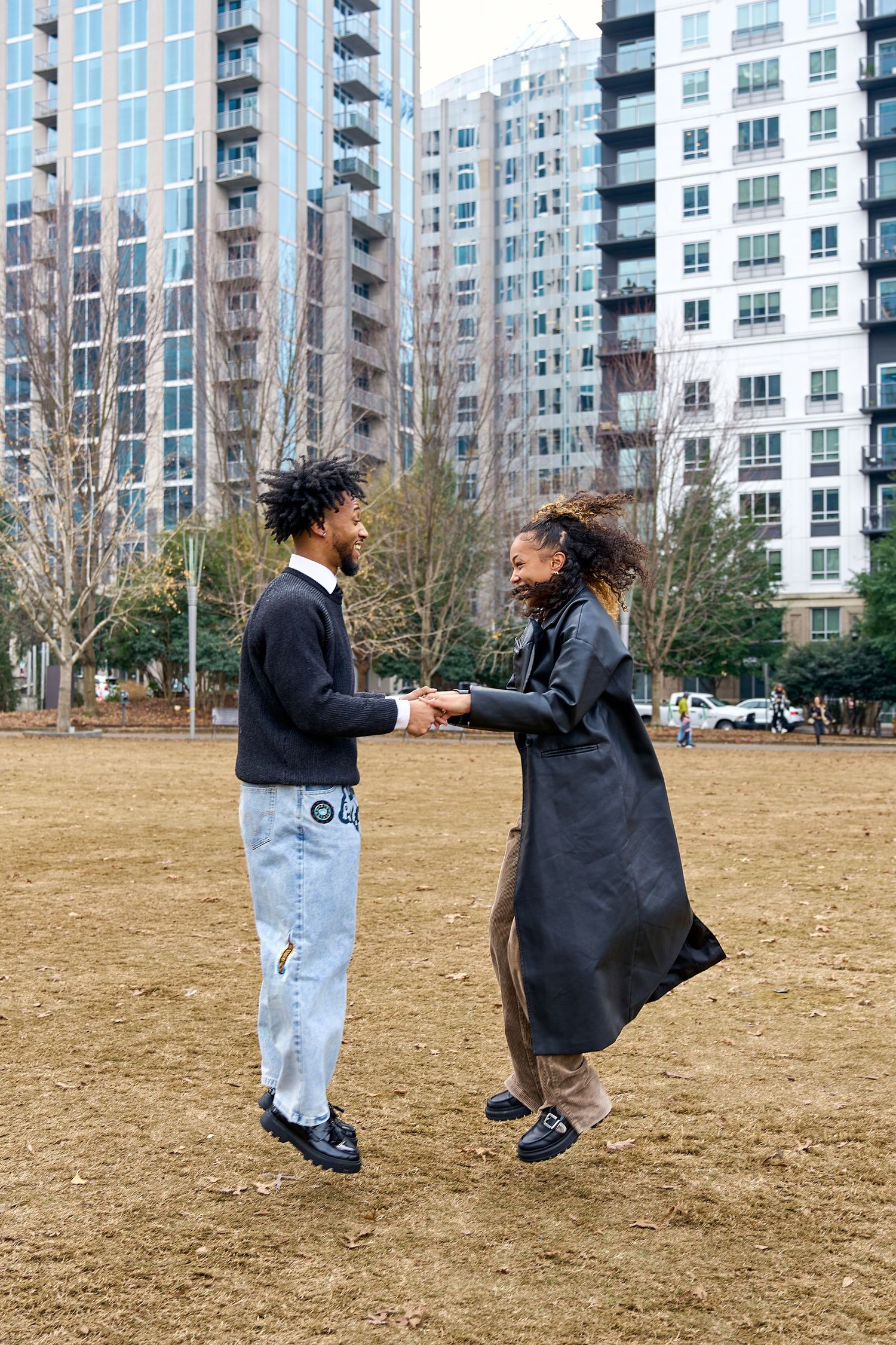 Two young people dancing and holding hands in an urban park with modern high-rise buildings in the background.