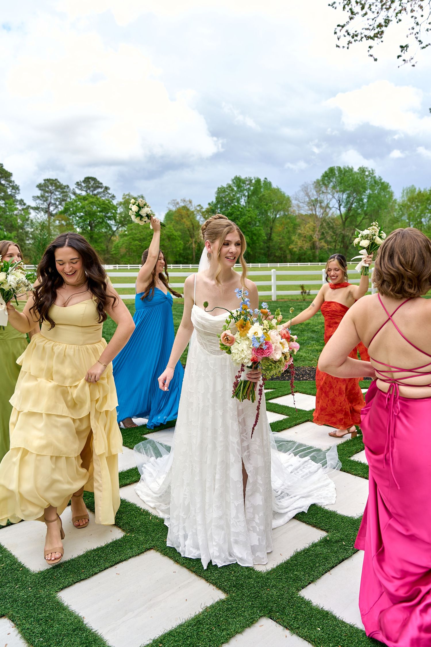 Bride in white lace gown walks with colorful bridesmaids in rainbow dresses on checkered aisle outdoors.