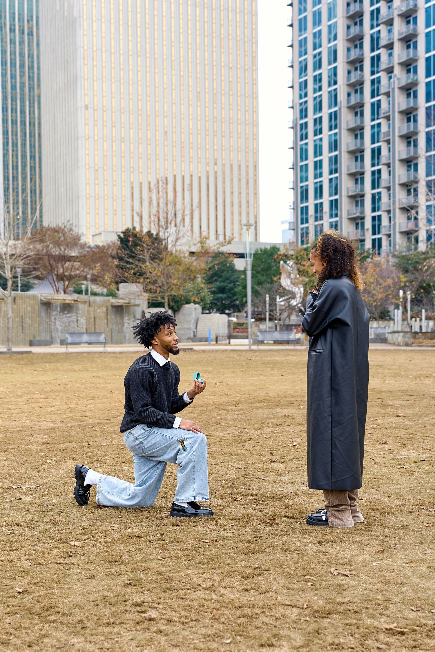 Man proposes on one knee to woman in urban park, holding ring box, with city skyscrapers in background.