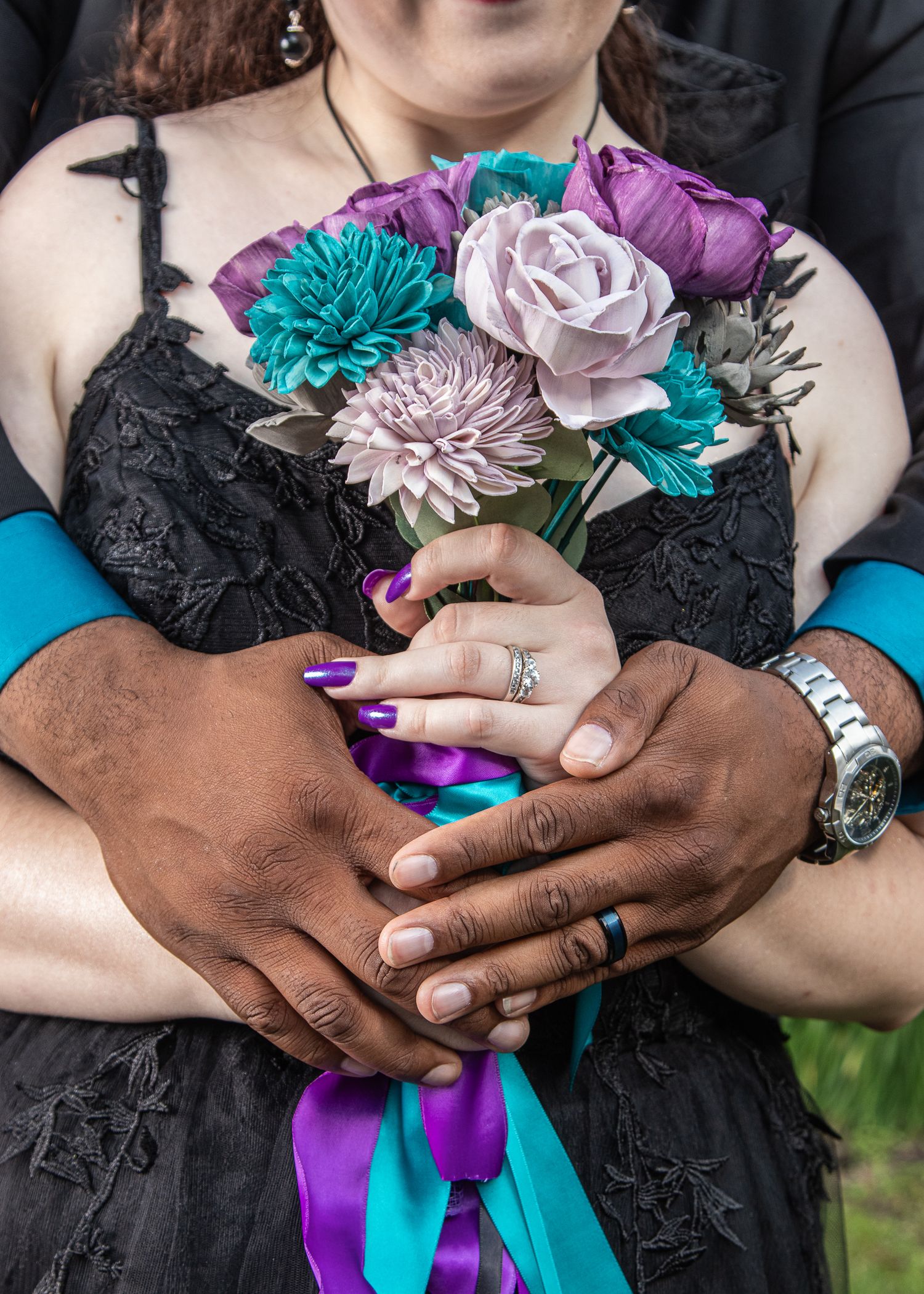 Interracial couple holding a bridal bouquet of purple and teal flowers, wearing wedding rings, dressed in black attire.