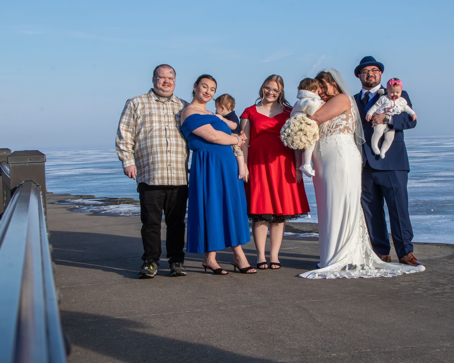 Wedding party poses outdoors on a waterfront boardwalk, bride in white gown with guests in red and blue attire, icy water behind.