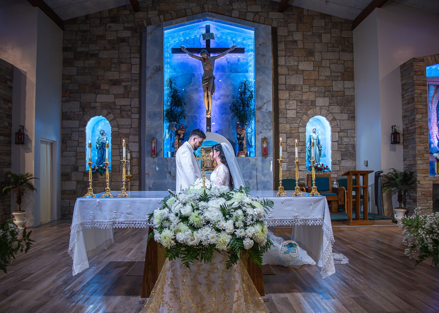 Couple kissing at church altar adorned with white floral arrangements during a Catholic wedding ceremony.