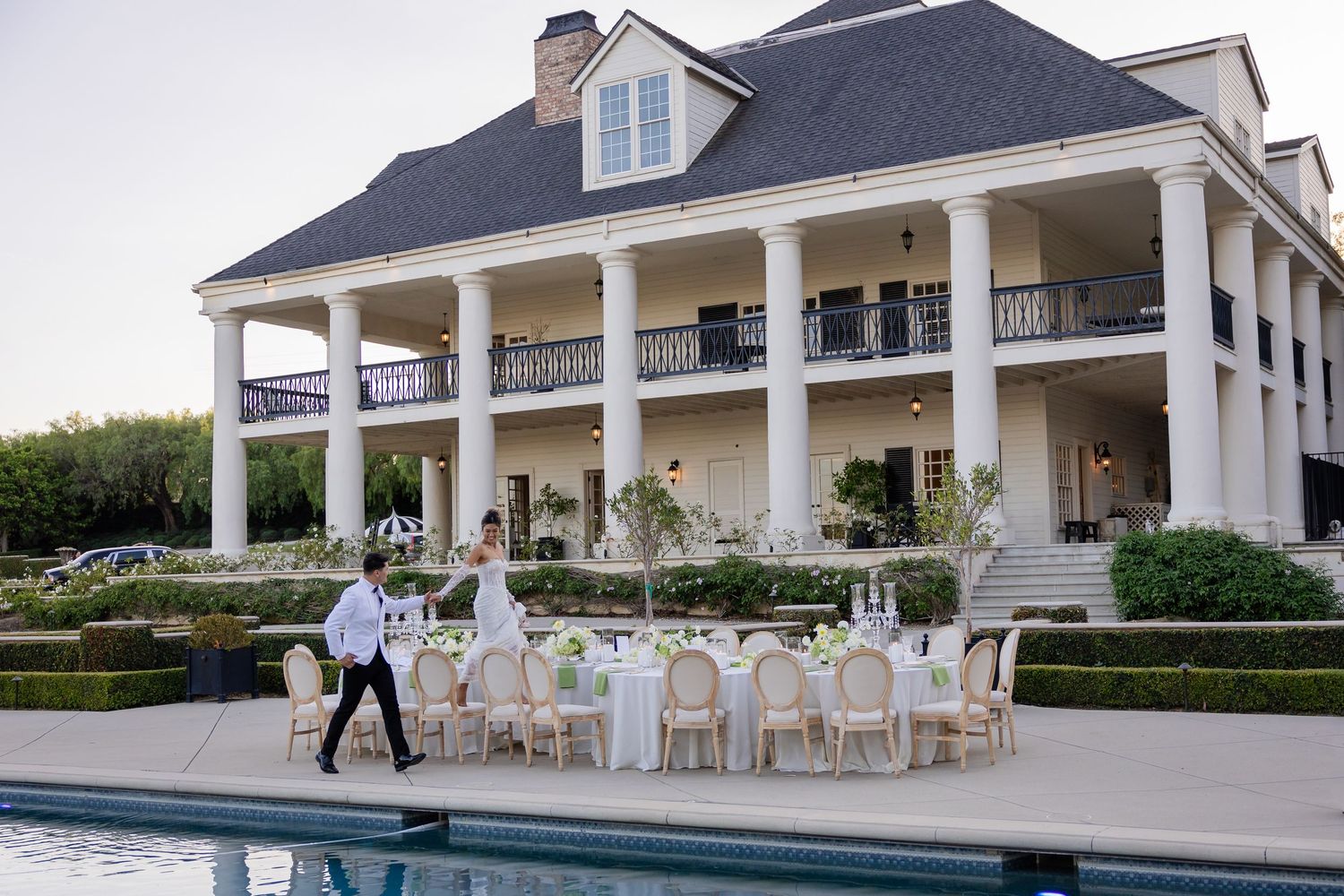 Waiter walking past elegant outdoor wedding reception tables beside a pool at a grand white-columned Southern estate.