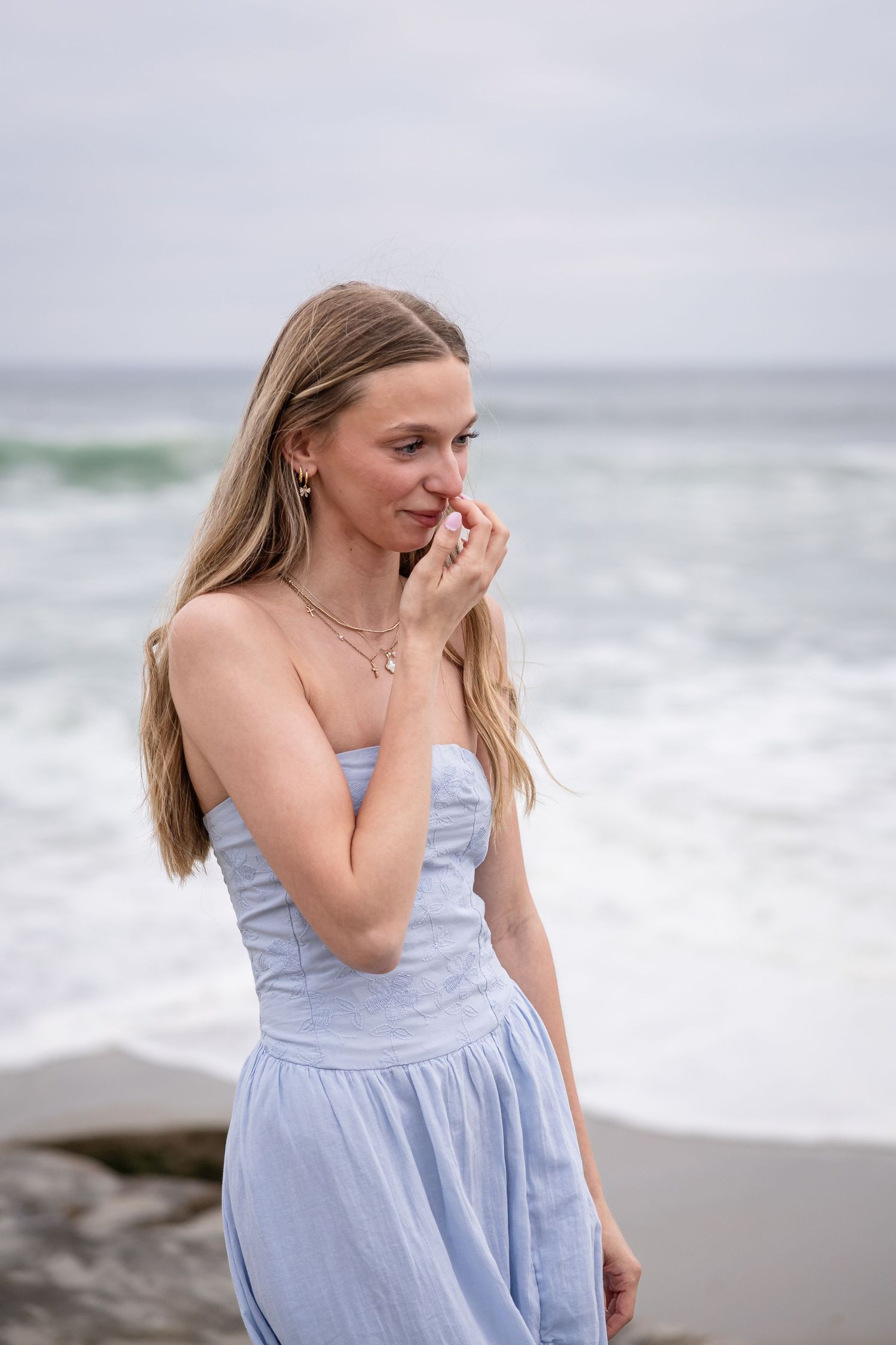 Young woman in light blue strapless dress standing on rocky beach with ocean waves in background.