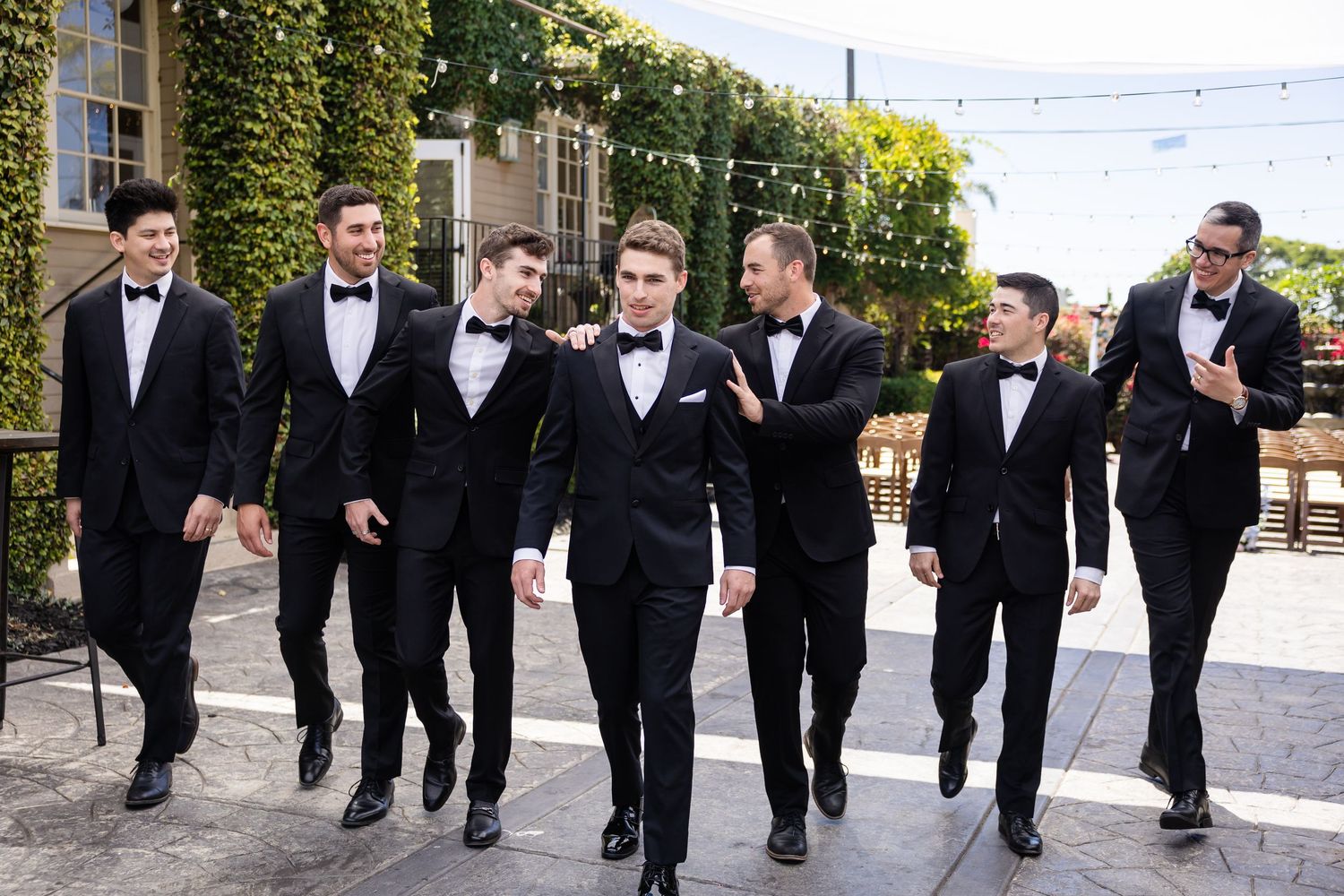 Seven groomsmen in black tuxedos and bow ties walking together outdoors at an elegant wedding venue.