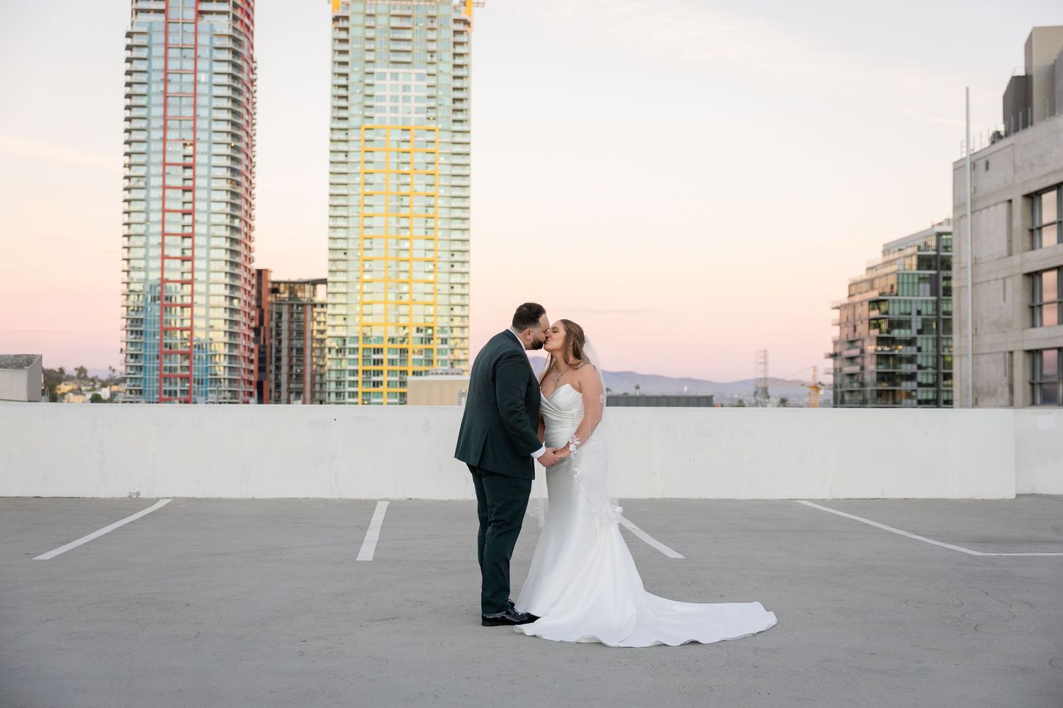 Bride and groom kissing on rooftop parking lot at sunset with city skyline skyscrapers in background.