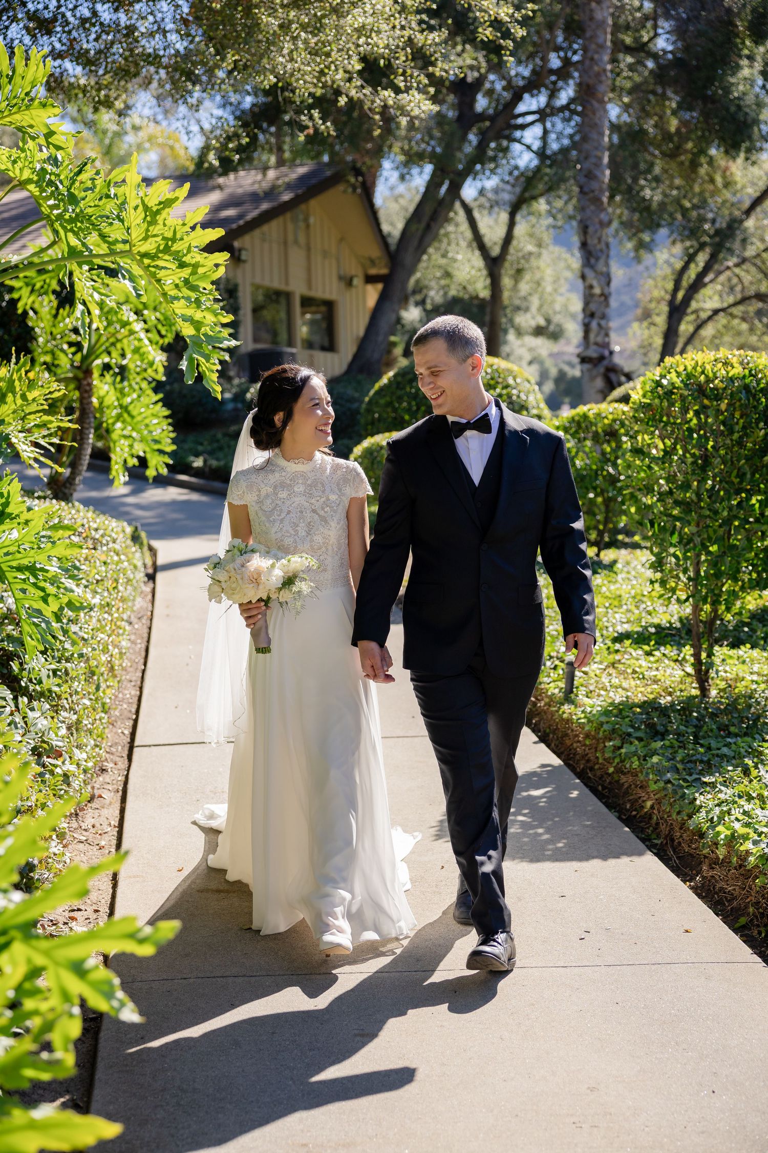 Bride holding bouquet and groom in black tuxedo walk hand in hand along a sunny garden pathway.