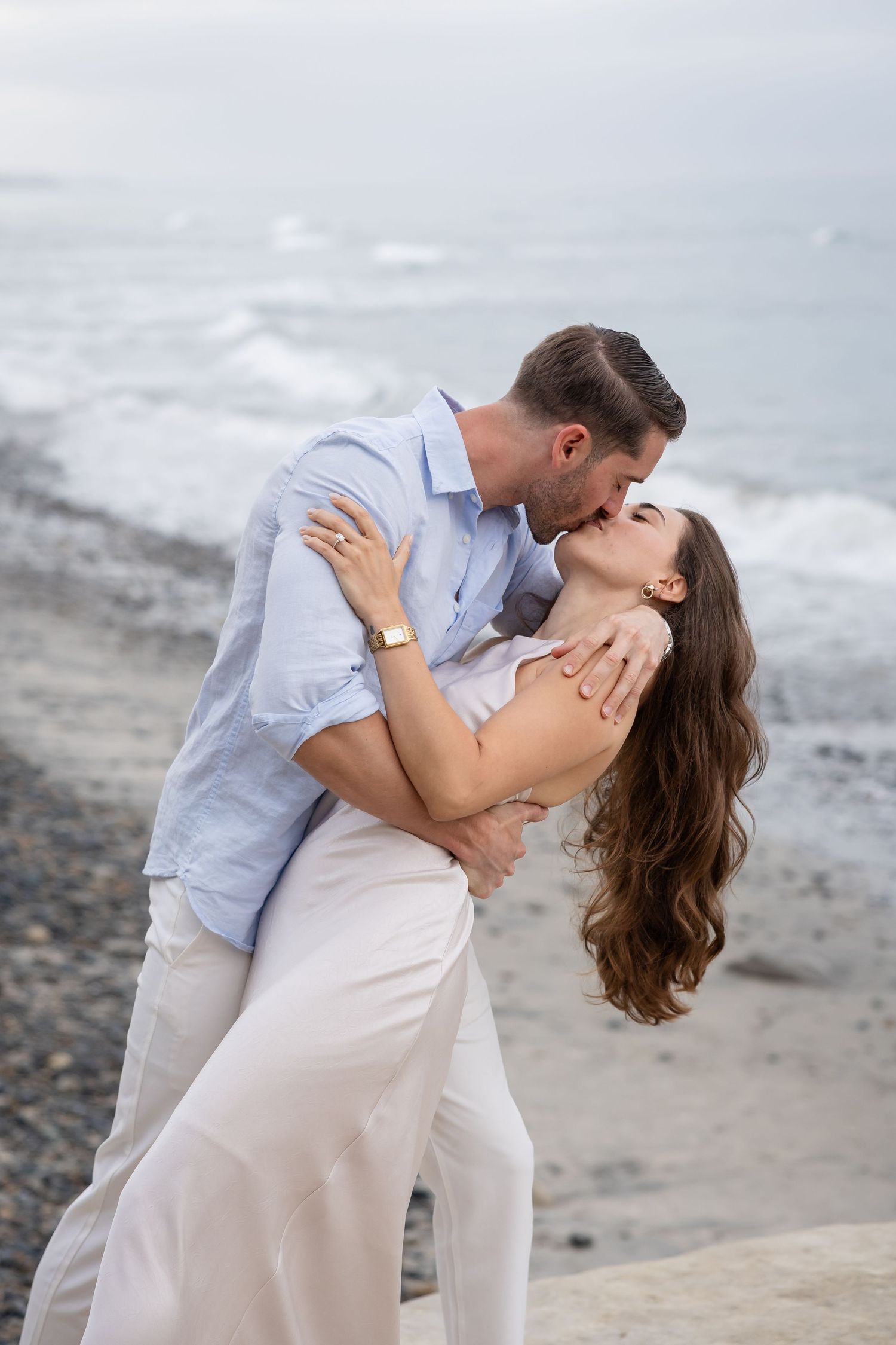 Romantic couple sharing a passionate kiss on a rocky beach, man dipping woman in white outfit by the ocean.