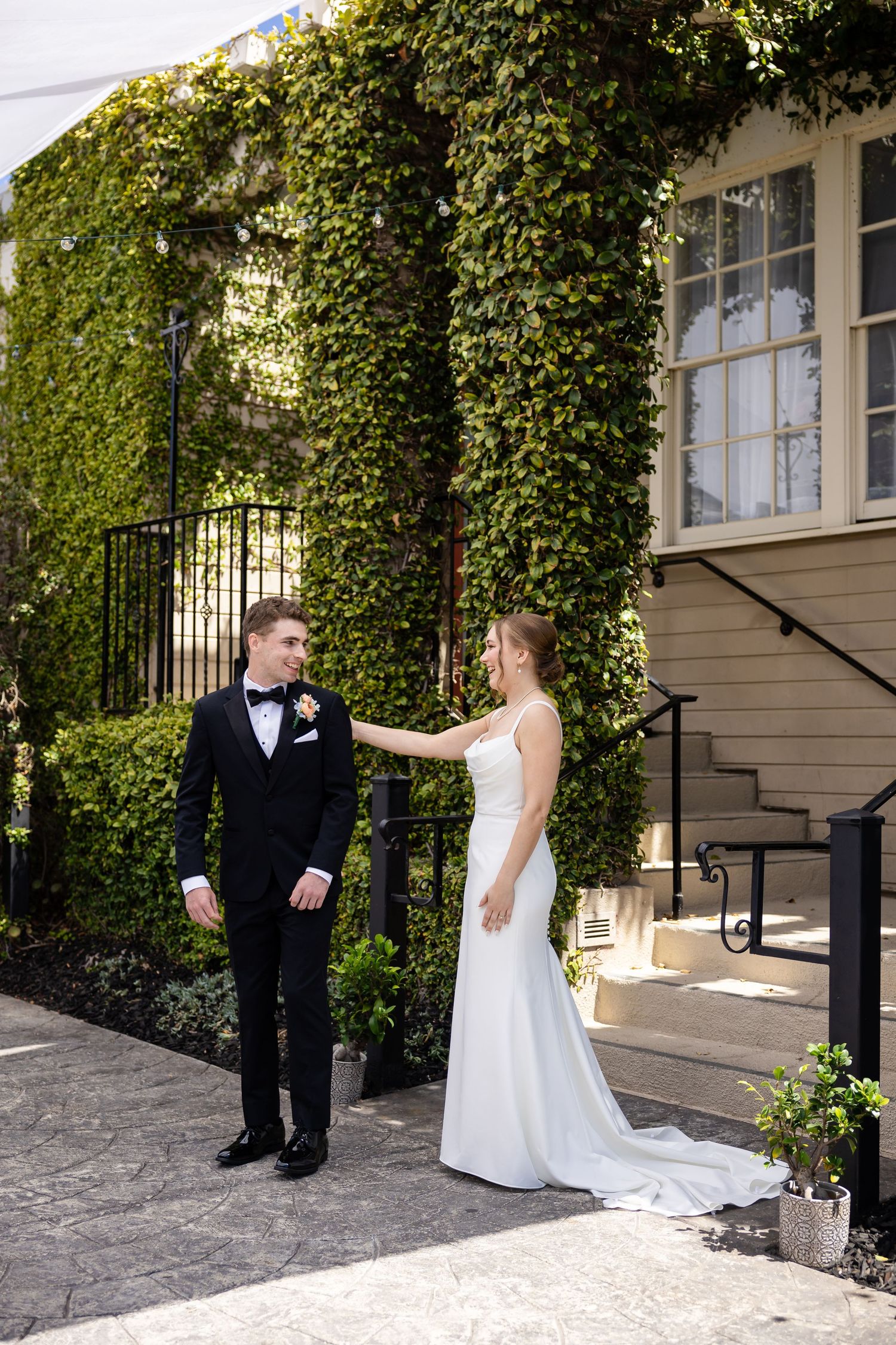 Bride in elegant white gown reaches out to groom in black tuxedo outside ivy-covered wedding venue.