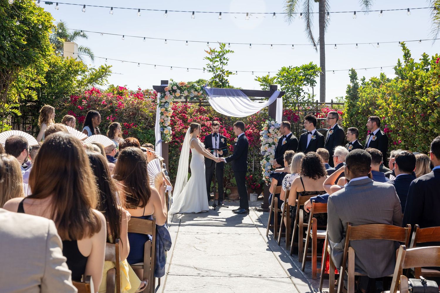 Outdoor wedding ceremony with bride and groom at floral arch, guests seated on wooden chairs under string lights.