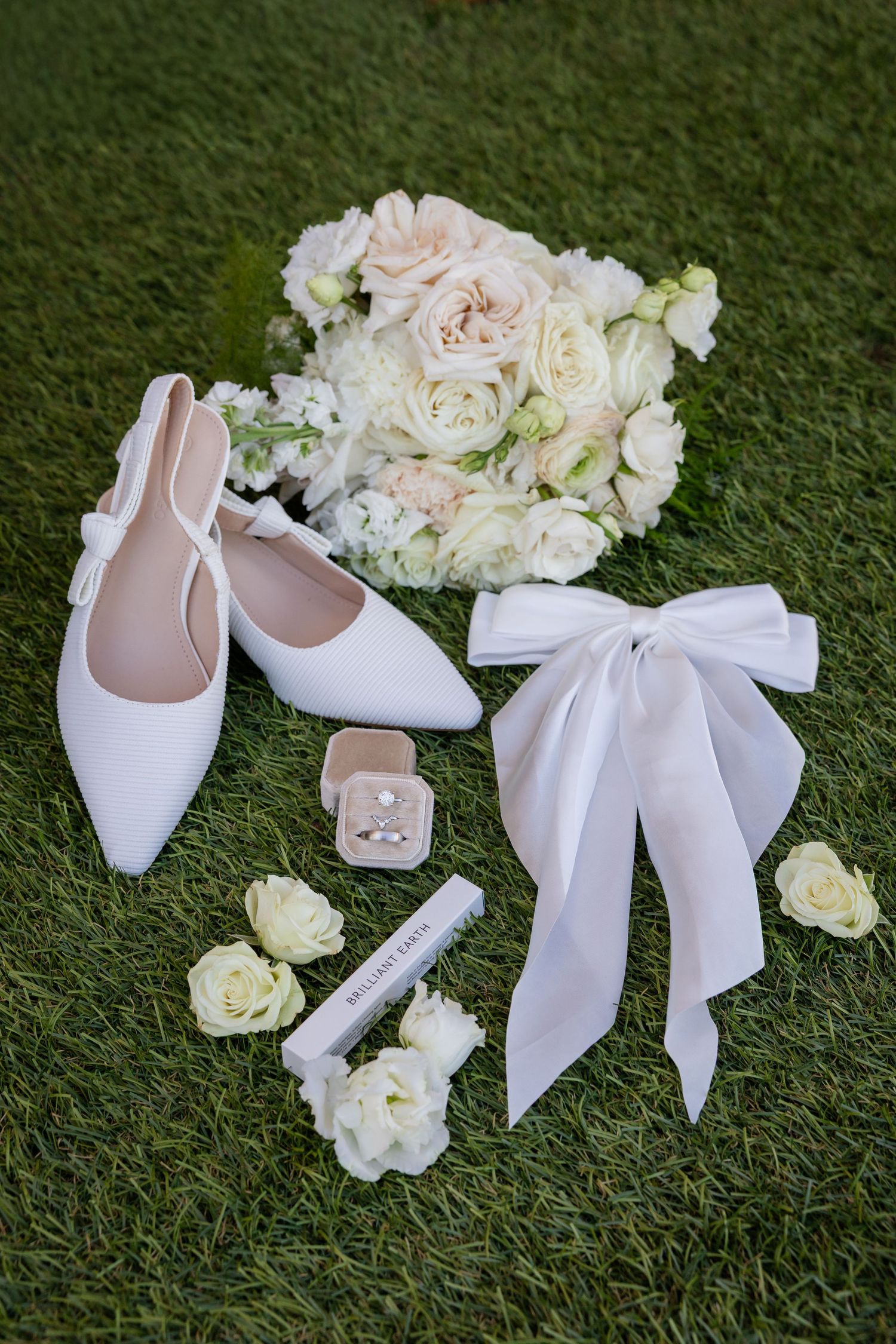 White bridal heels, bouquet, ring box, and ribbon bow accessories arranged on green grass for wedding flat lay.