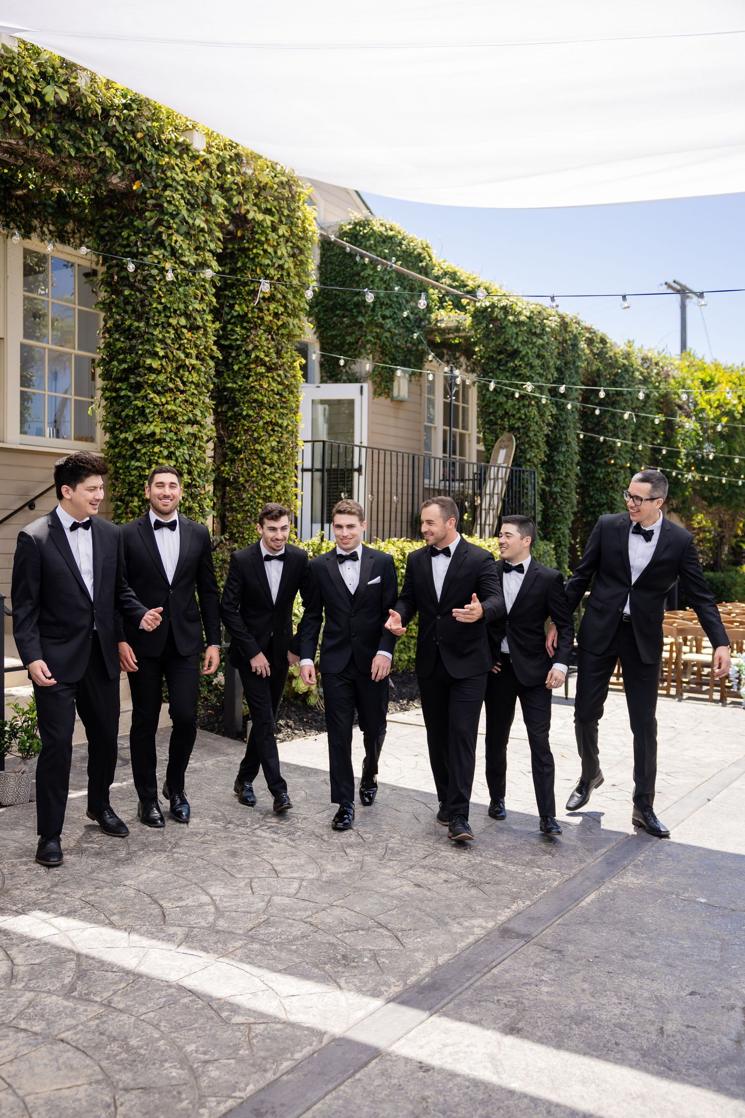 Six groomsmen in matching black tuxedos walk together laughing at an ivy-covered wedding venue.