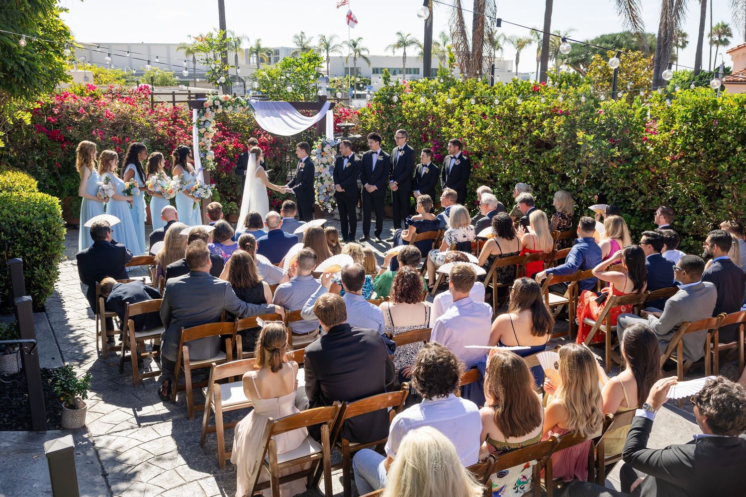 Outdoor wedding ceremony with guests seated on wooden benches surrounded by colorful flowers and palm trees in a garden setting.