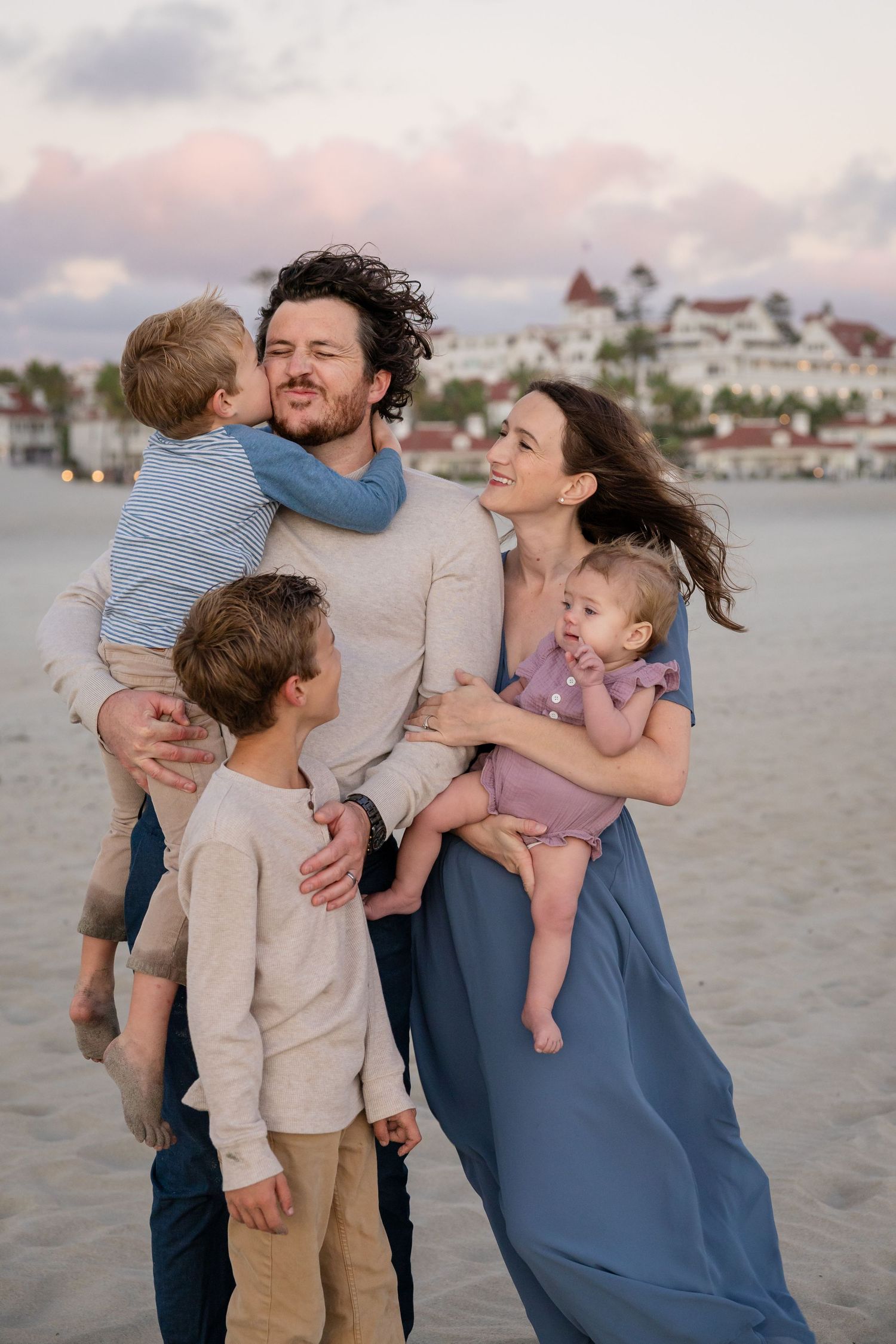 Family of five poses on a beach at sunset, parents holding young children with a historic hotel in the background.