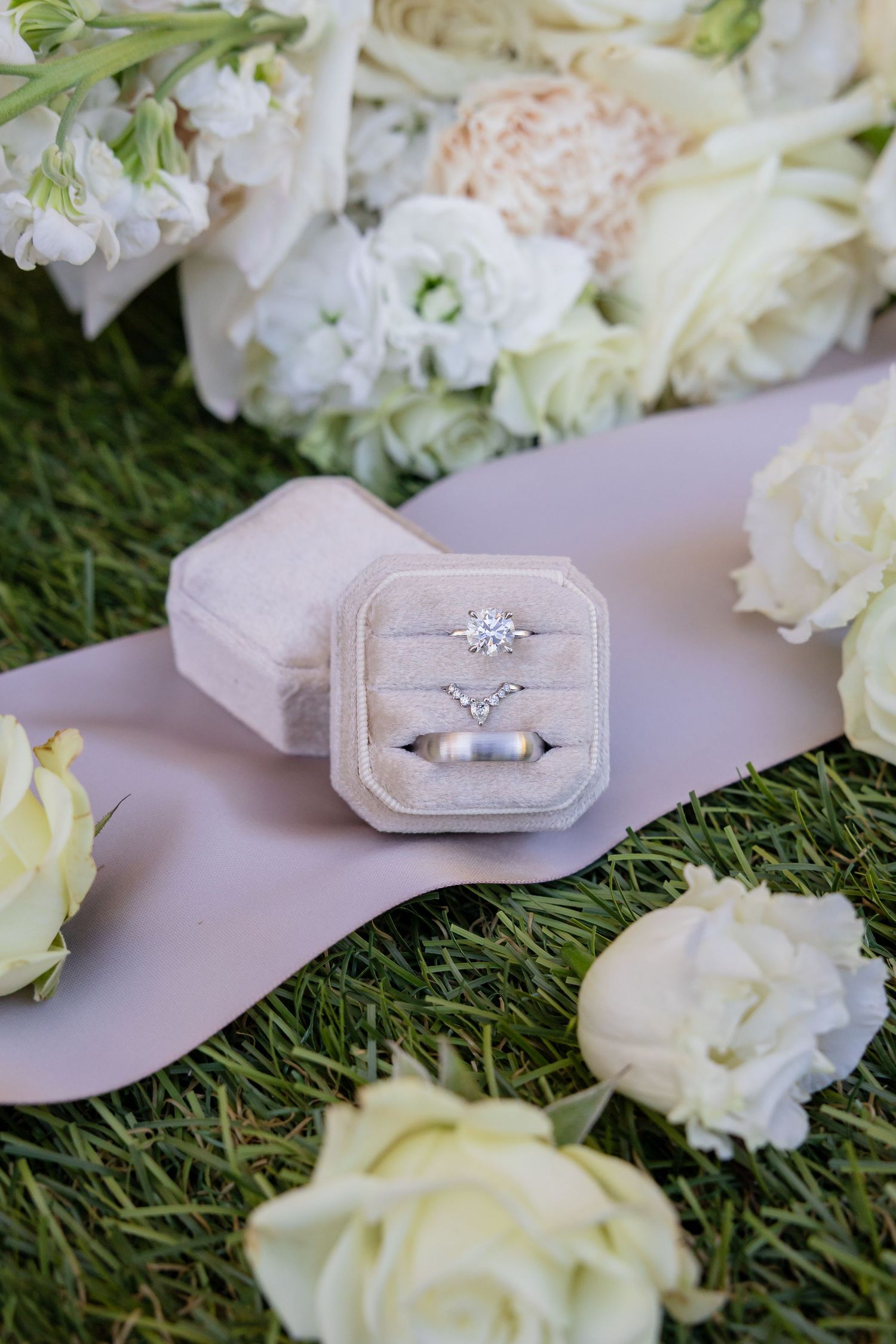 Diamond engagement ring and wedding band displayed in velvet ring box surrounded by white roses on grass.