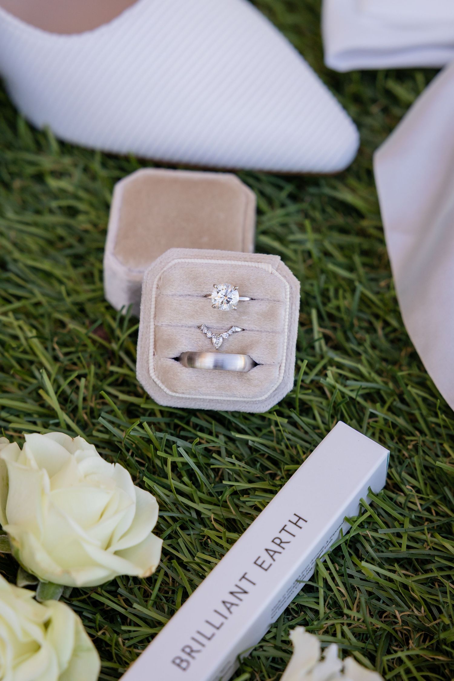 Brilliant Earth diamond engagement ring and wedding bands displayed in a velvet box on green grass with white roses and heels.