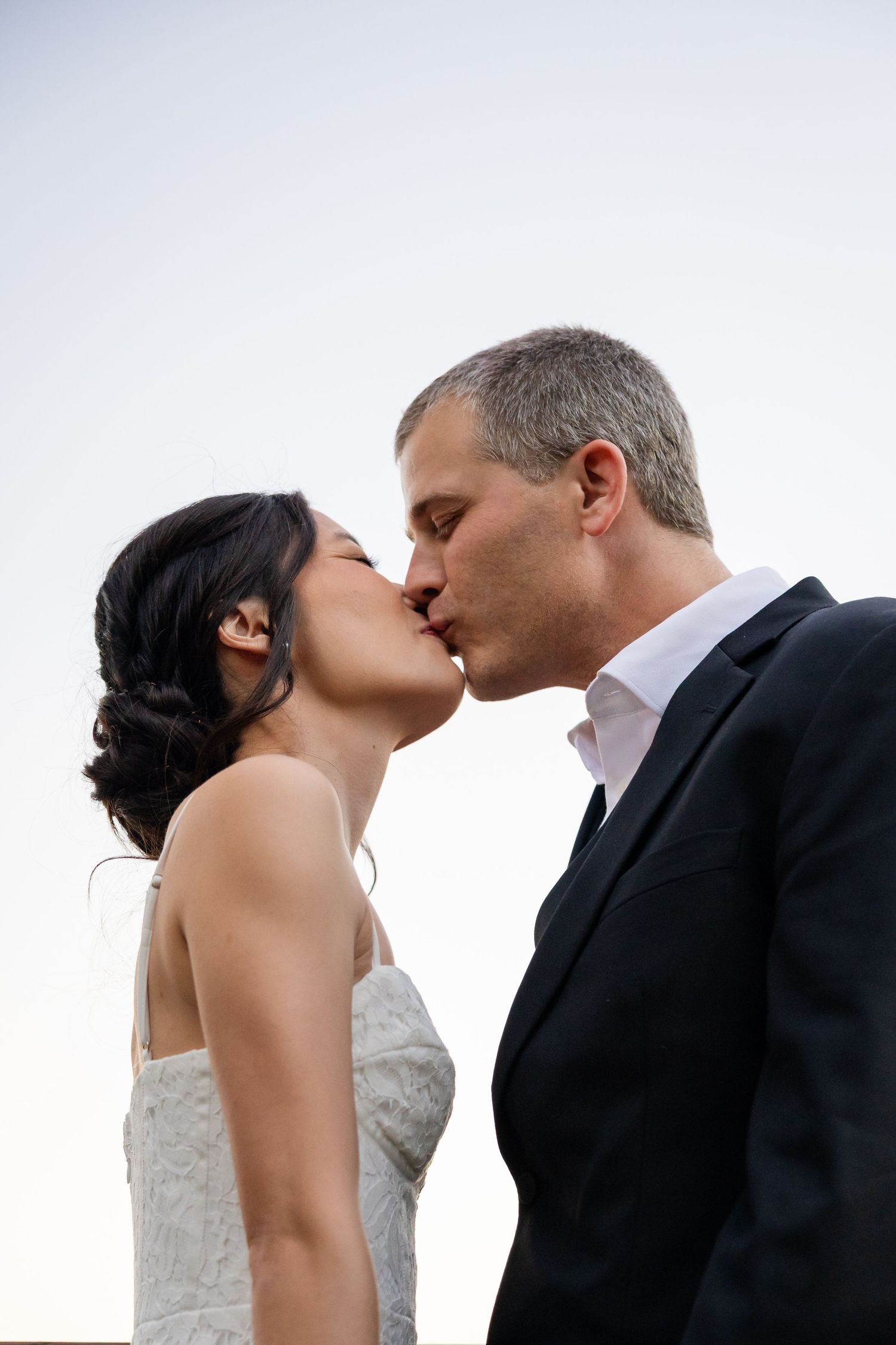 Bride and groom share a romantic kiss against a soft blue sky in their wedding attire.