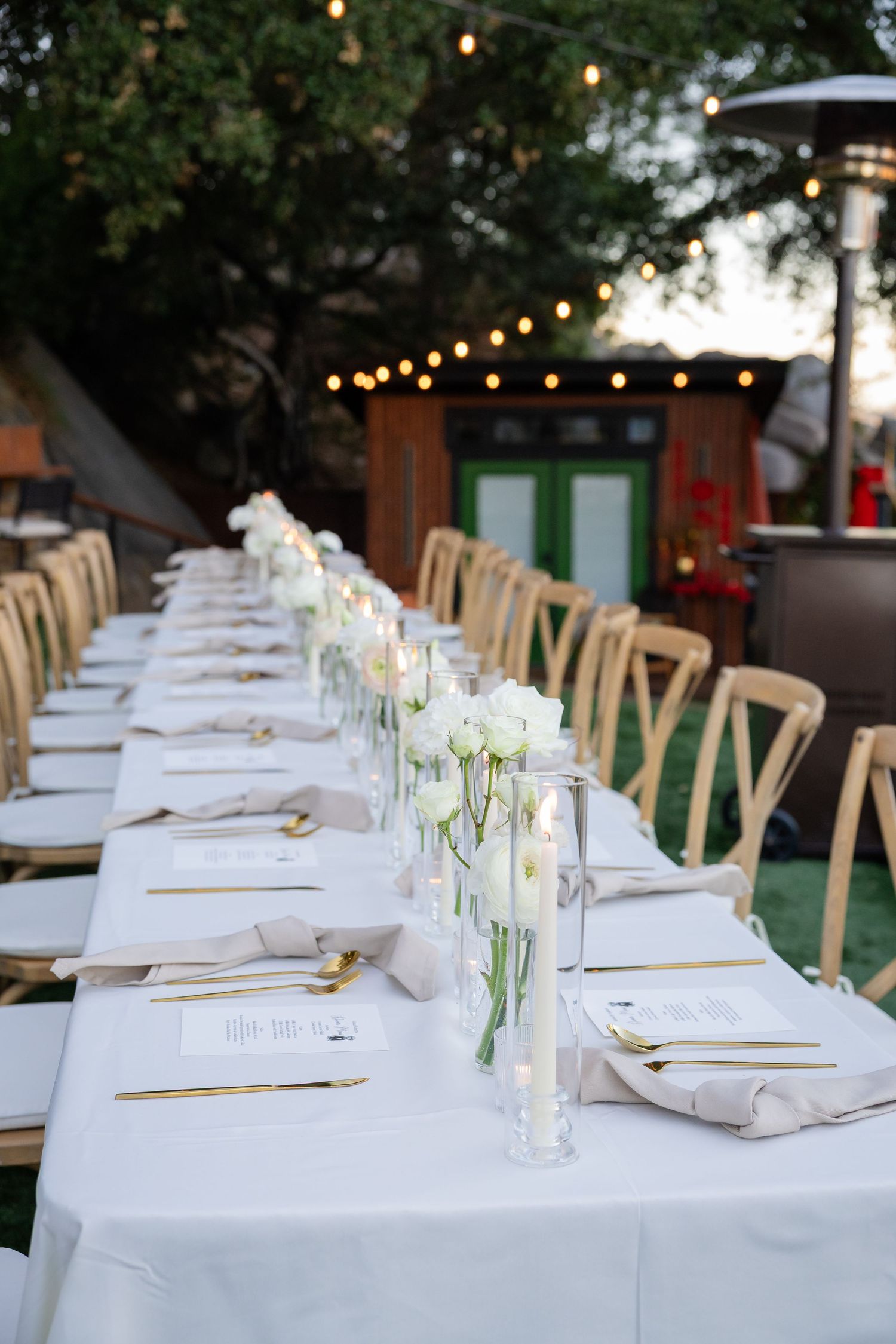 Elegant outdoor wedding reception table set with white linens, white ranunculus, candles, and gold accents.