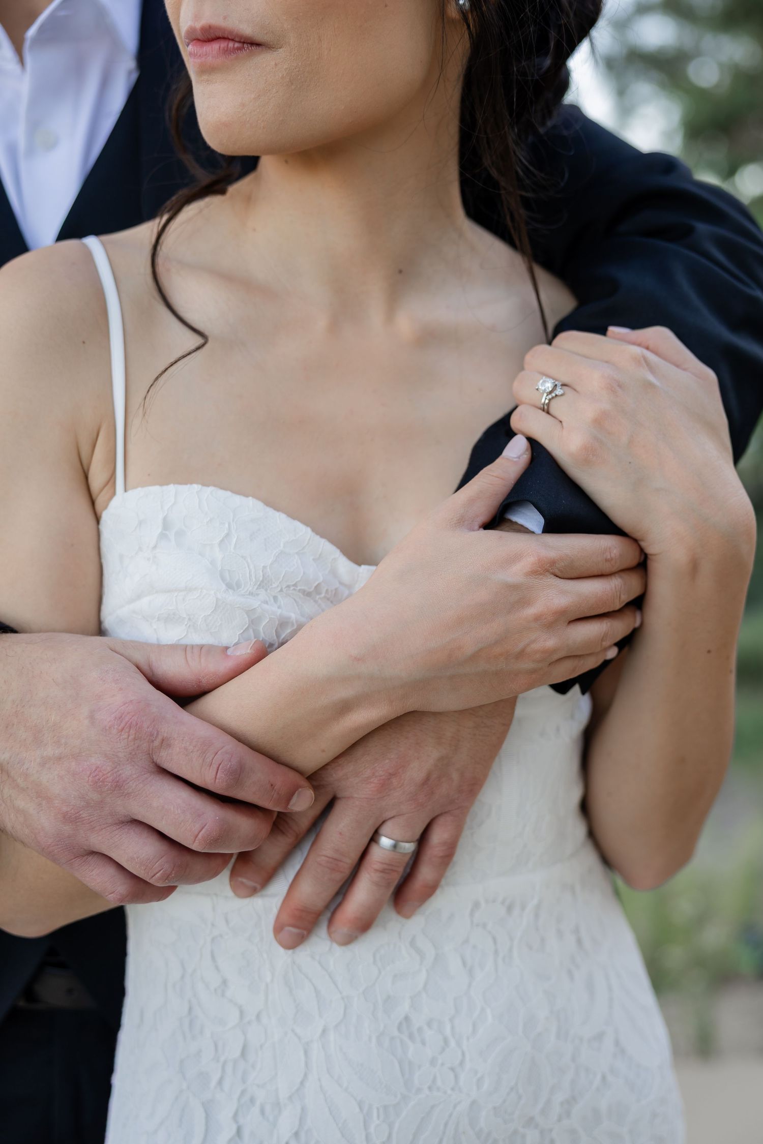 Close-up of bride in lace gown and groom embracing, showcasing diamond engagement ring and wedding band.