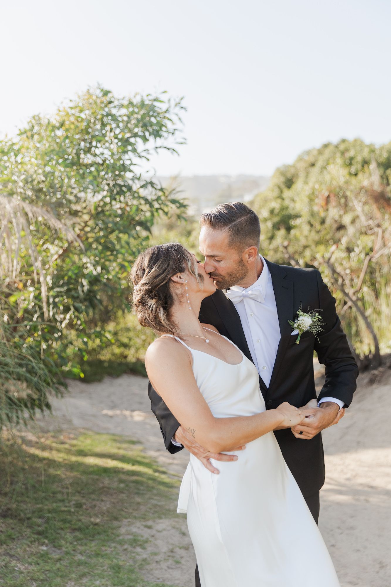 A bride and groom share a tender kiss outdoors on a sunlit dirt path surrounded by lush green foliage.