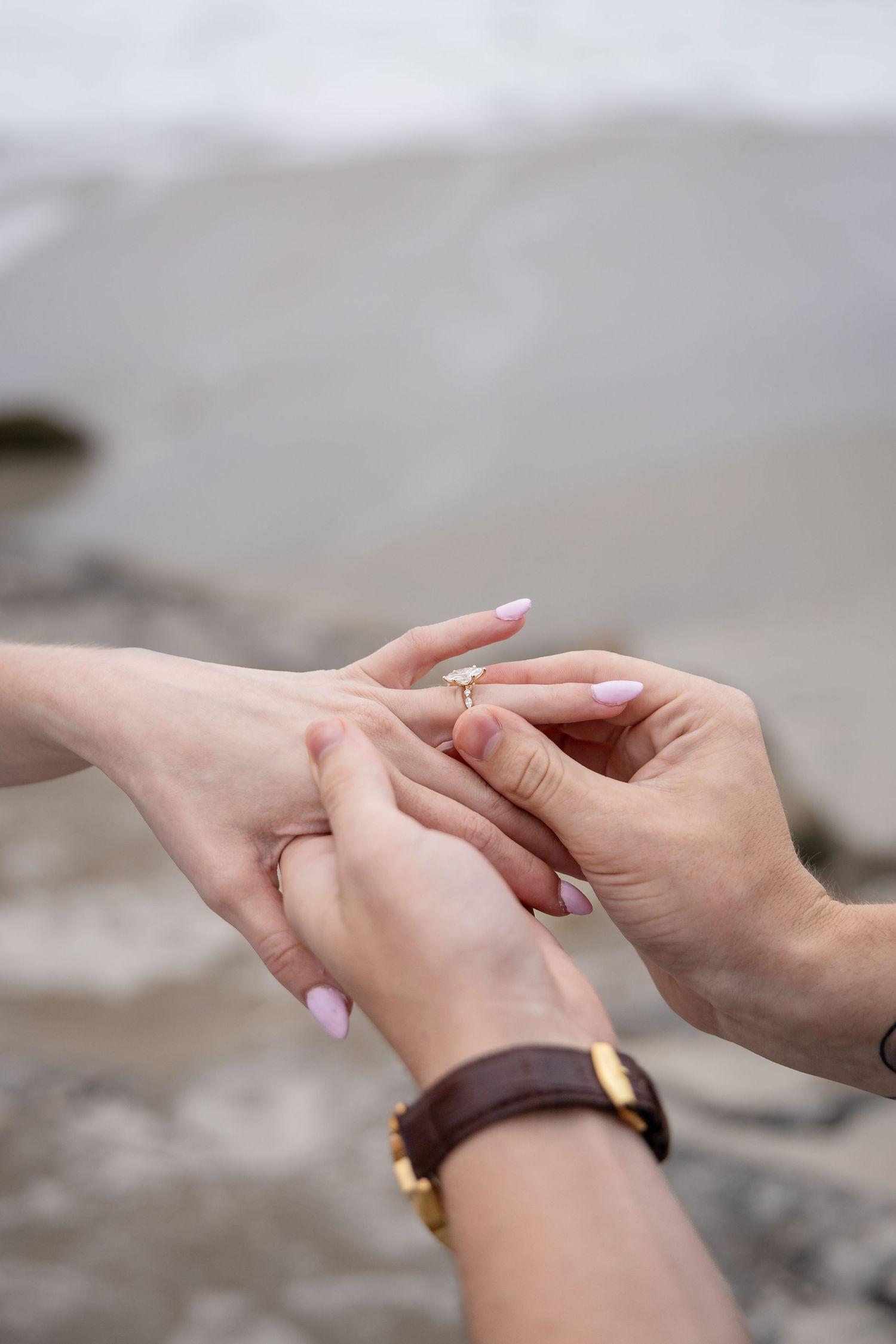 Person placing a gold diamond engagement ring on a woman's finger outdoors near a rocky beach.