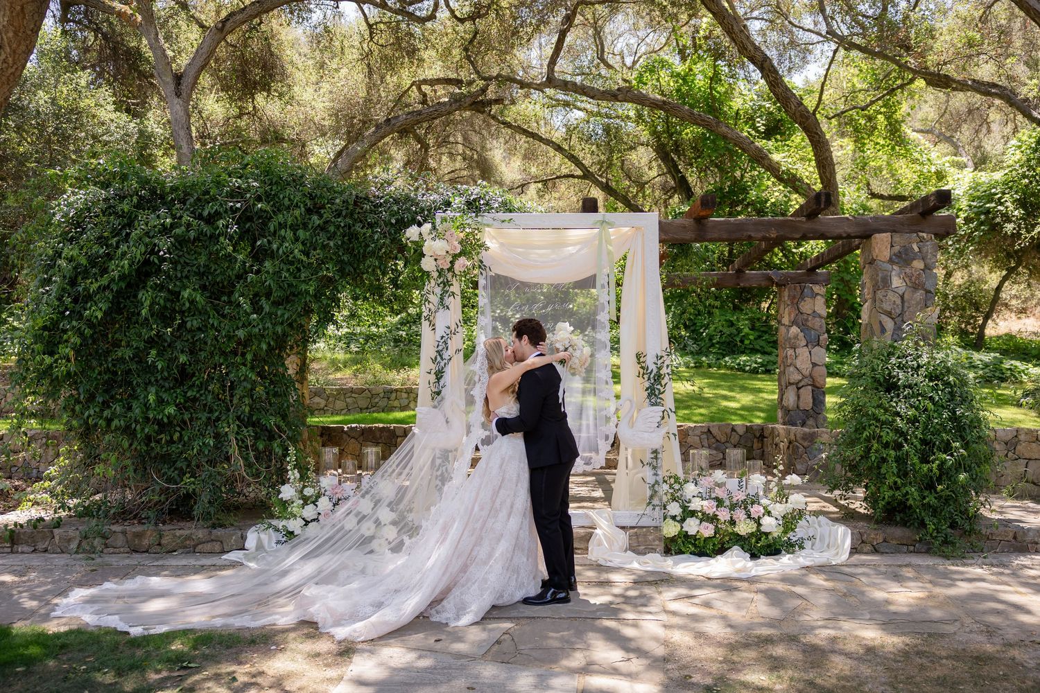 Bride and groom share first kiss under floral arch surrounded by lush greenery at outdoor garden wedding ceremony.