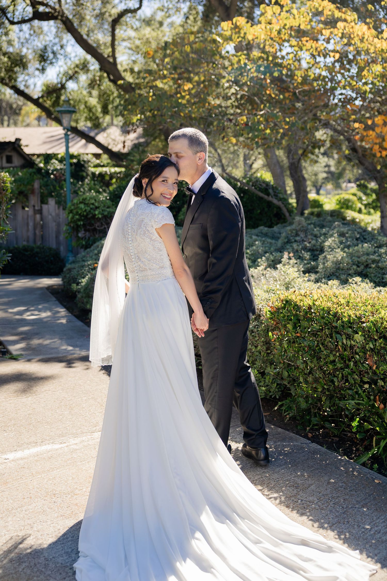 Bride in white gown with veil holds groom's hand while looking back smiling on a sunny garden path.