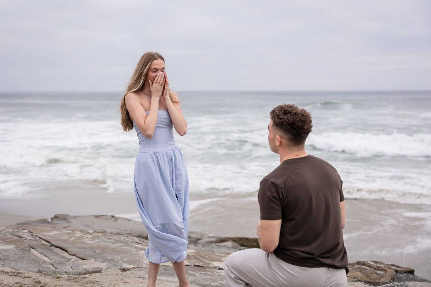 Man proposing to surprised woman on rocky beach with ocean waves in background.