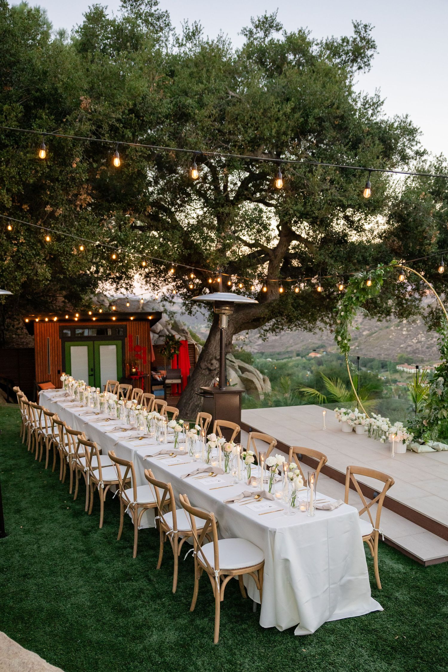 Elegant outdoor wedding reception with long white-draped tables, wooden cross-back chairs, string lights, and lush green trees at dusk.
